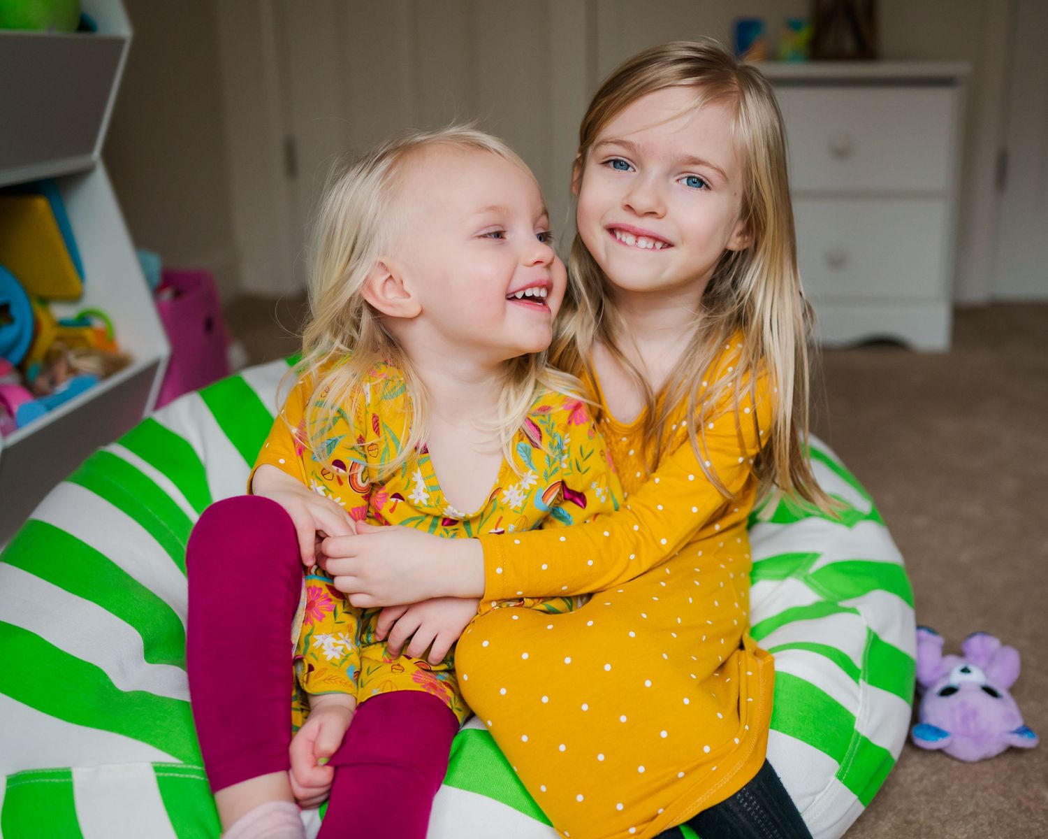 Two children wearing yellow floral dresses share a joyful moment on a green striped beanbag chair.