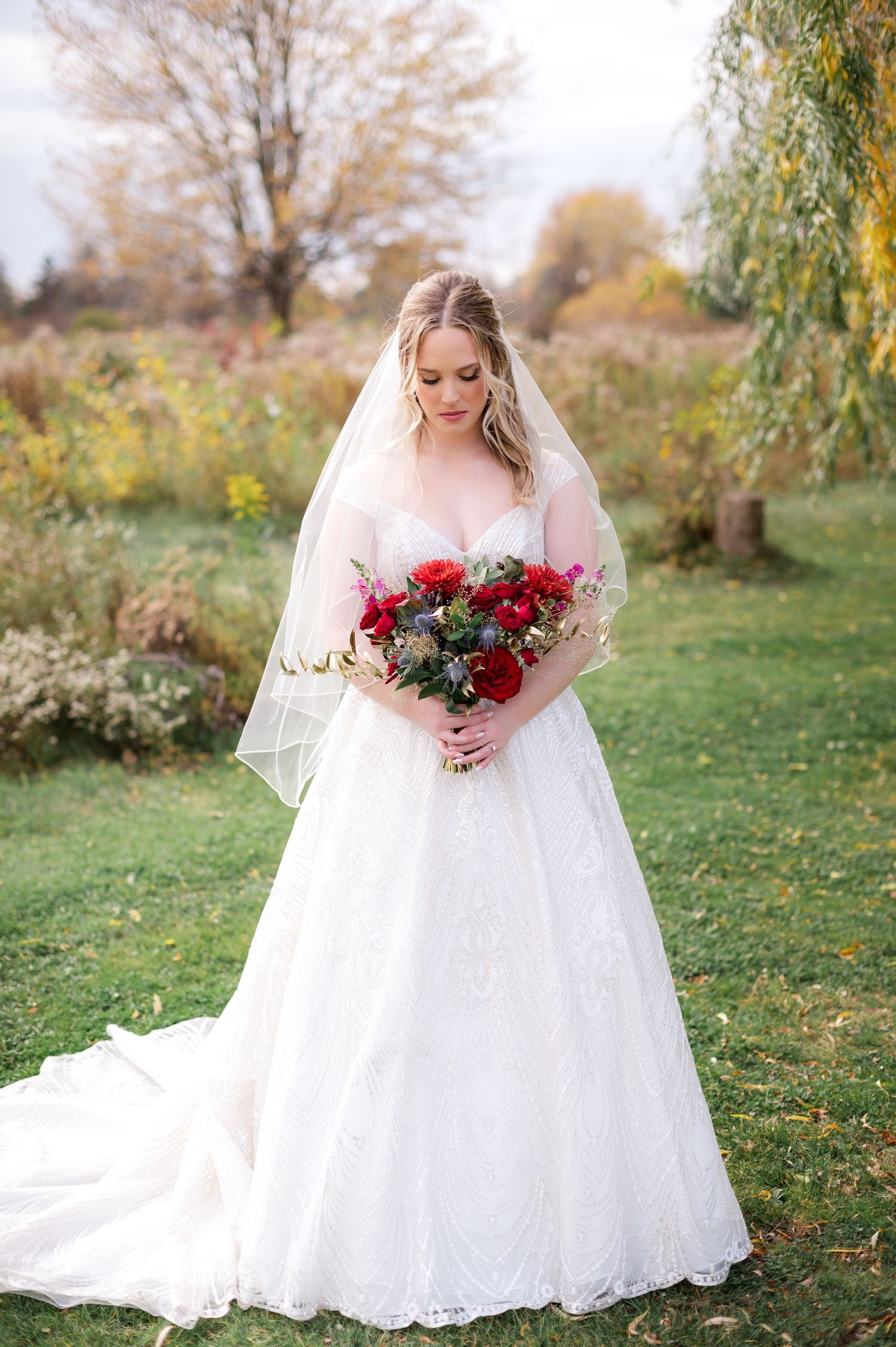 Bride in white lace gown holding red bouquet in garden setting during fall.