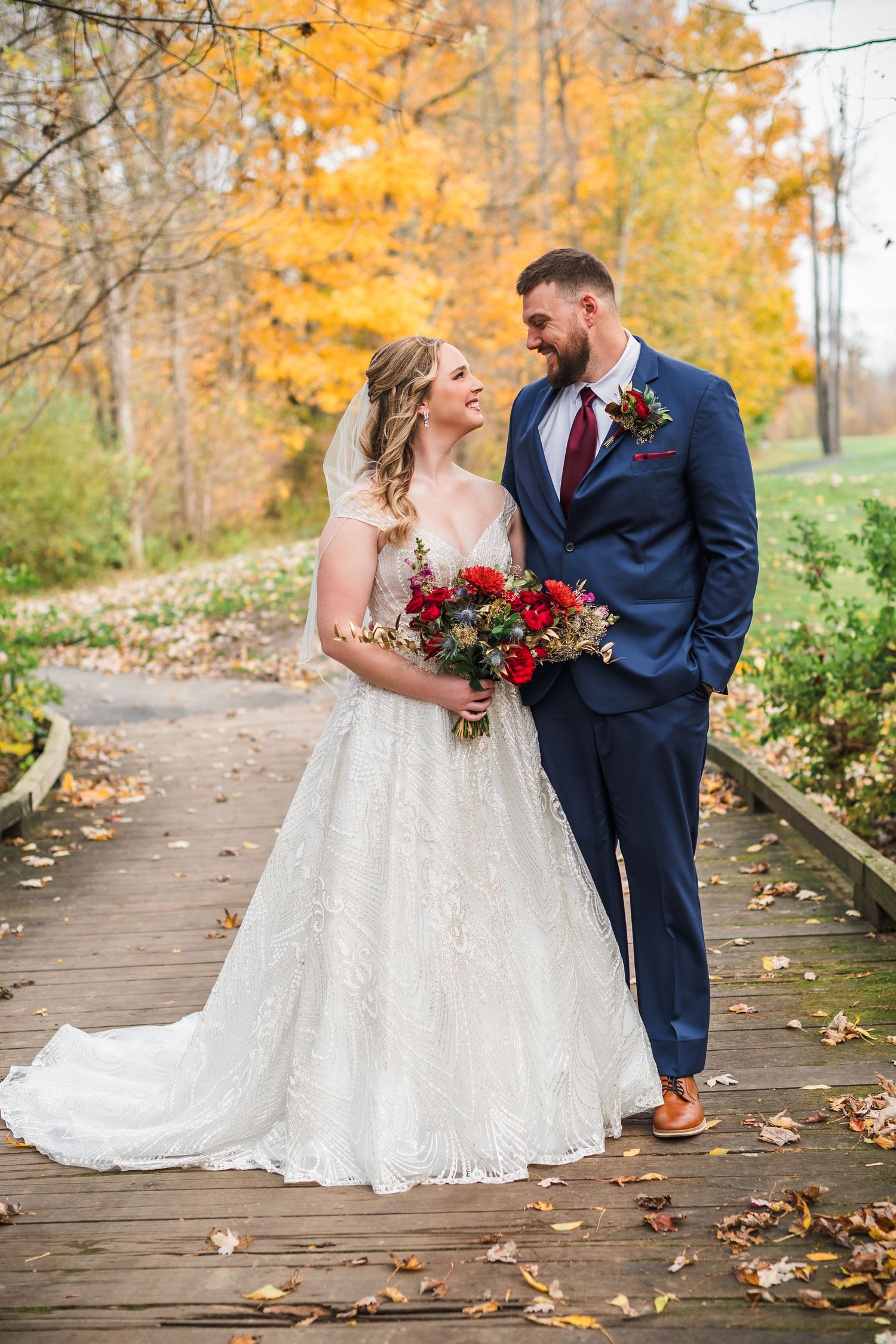 Couple poses on wooden walkway surrounded by vibrant fall colors and holding red bouquet.