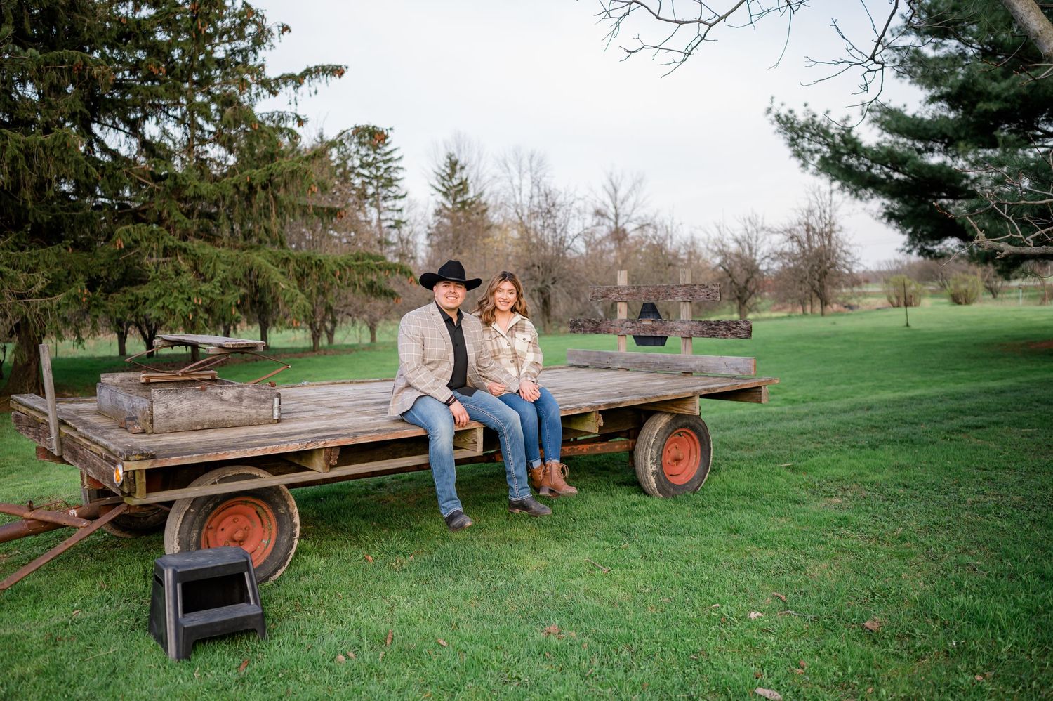 A couple sits together on a vintage farm wagon in a grassy field surrounded by trees.