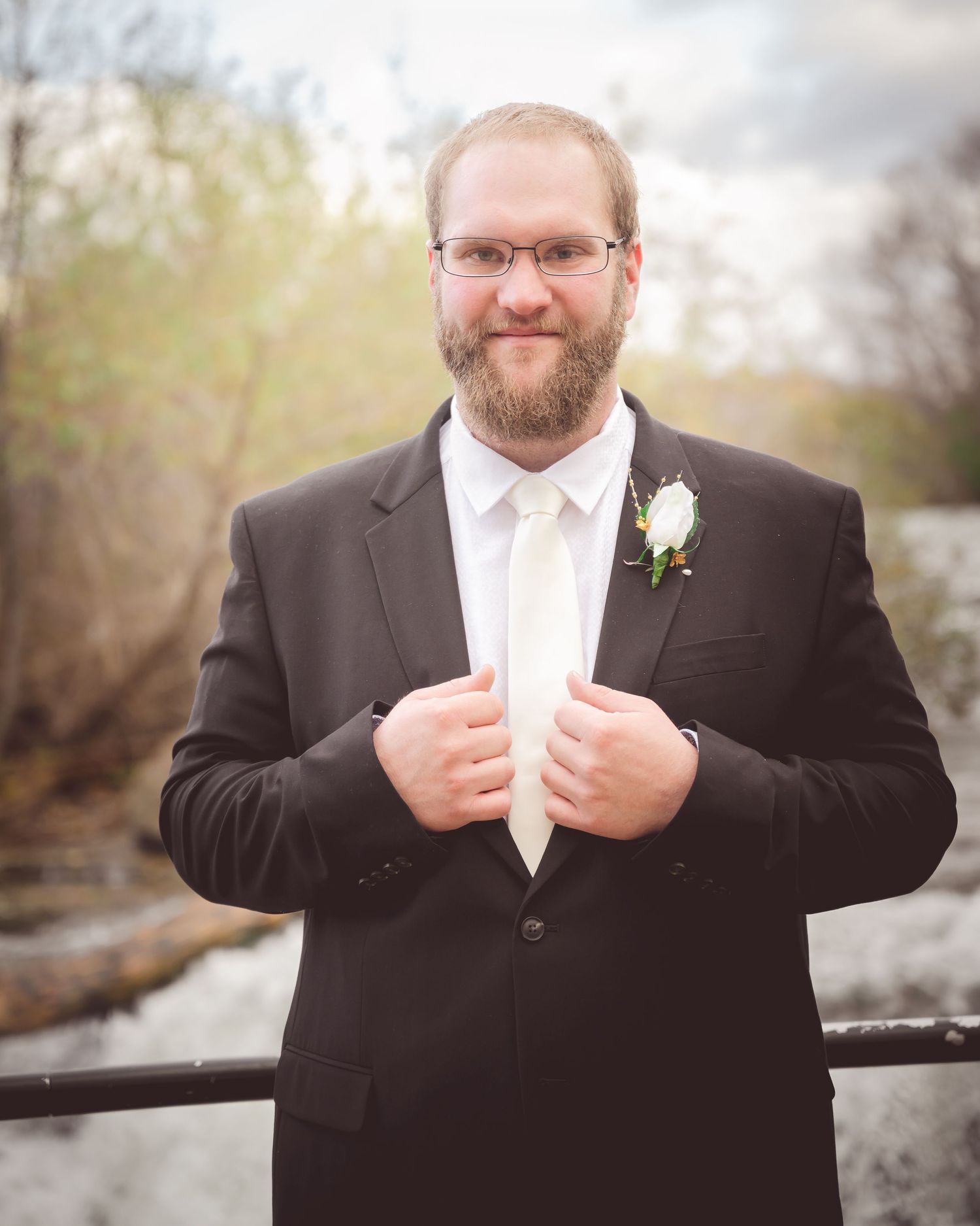 Person in formal black suit with white boutonniere standing outdoors.