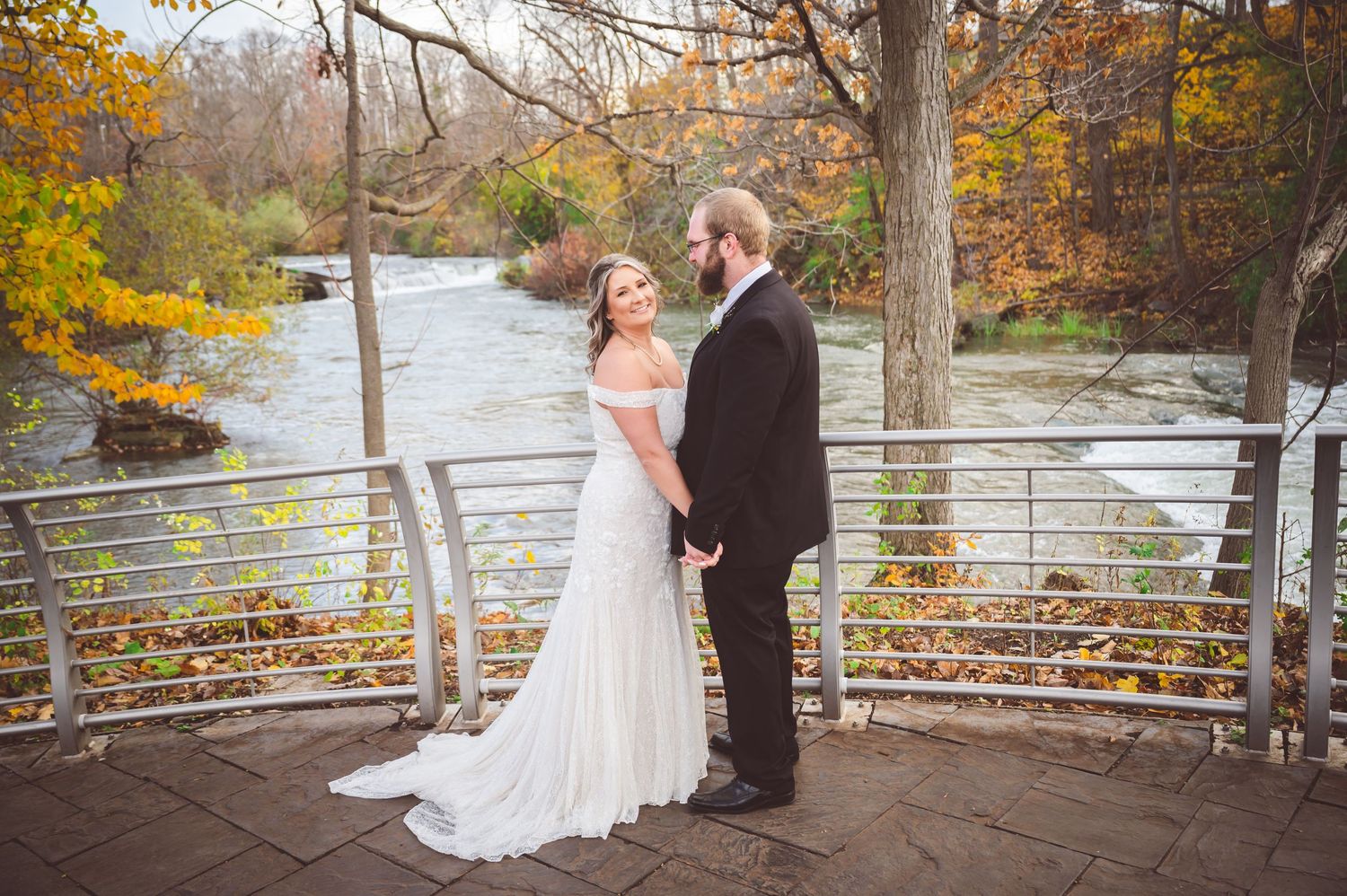 A couple stands together on a stone observation deck overlooking a river surrounded by fall colors.