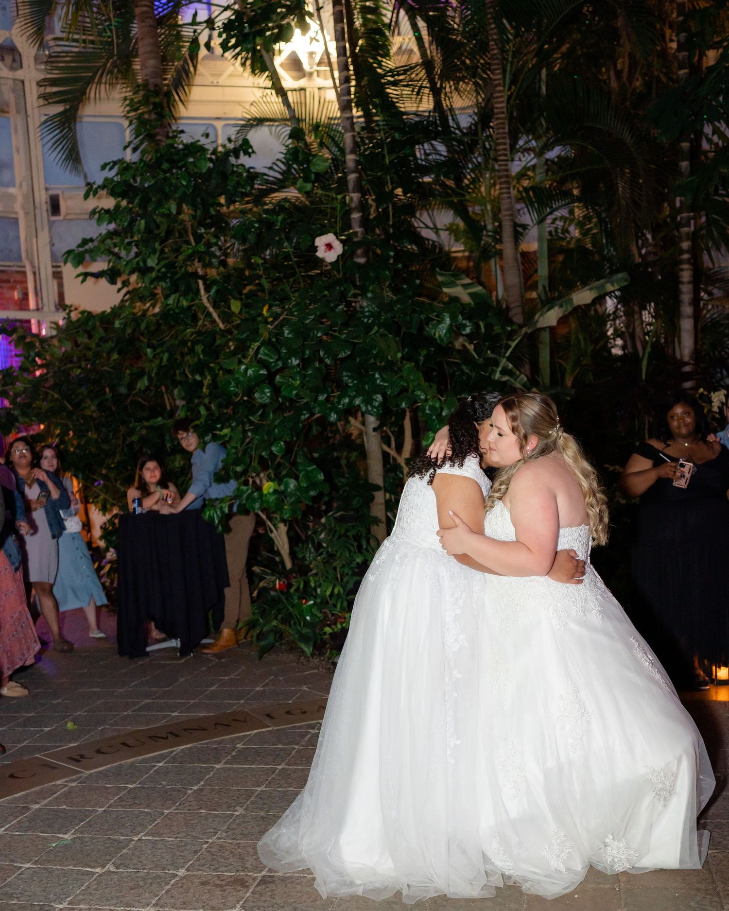 Two brides share a romantic dance surrounded by guests and garden foliage.