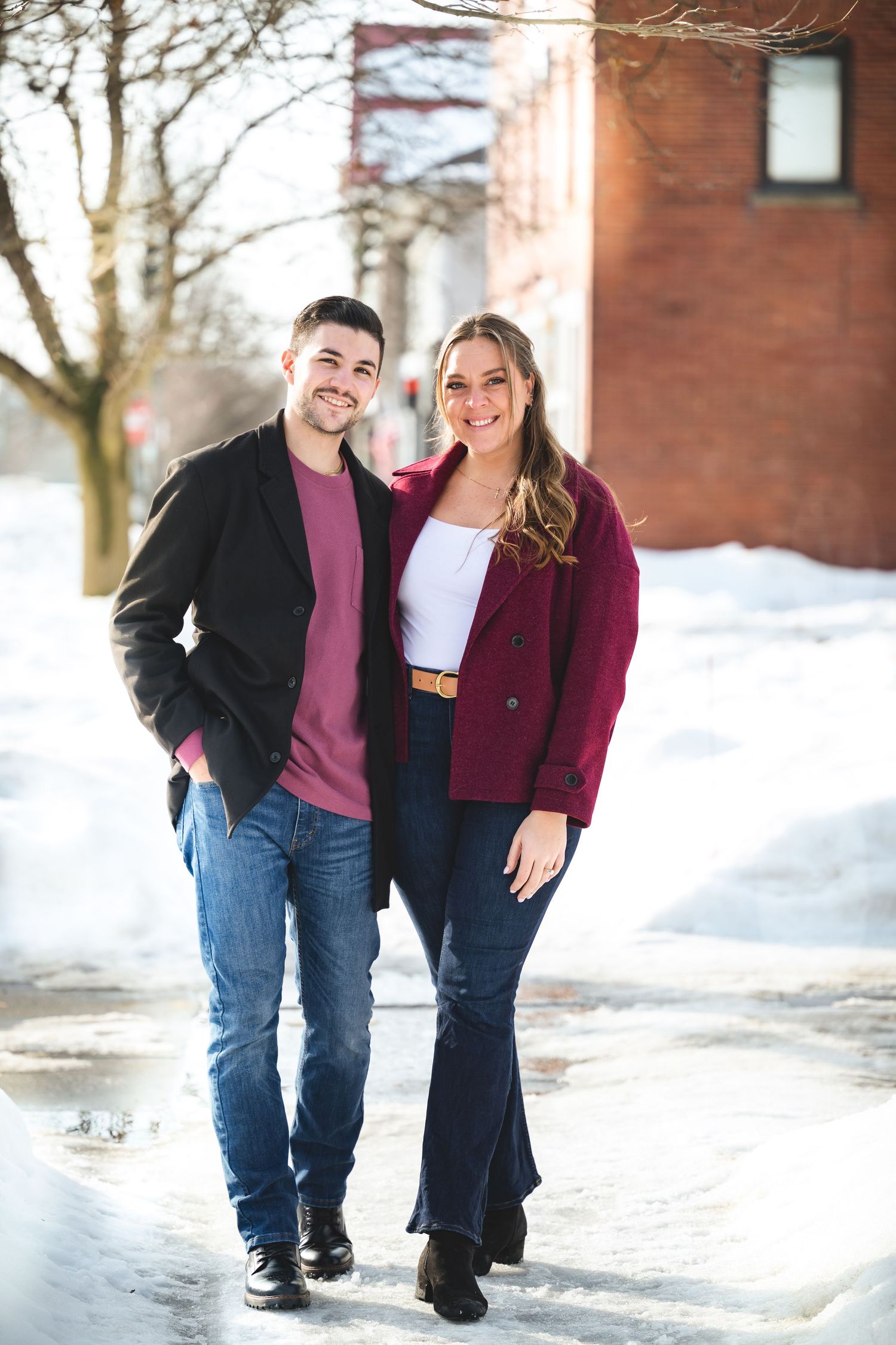 A couple poses together in the snow against a brick building wall wearing coordinating burgundy and black outfits.