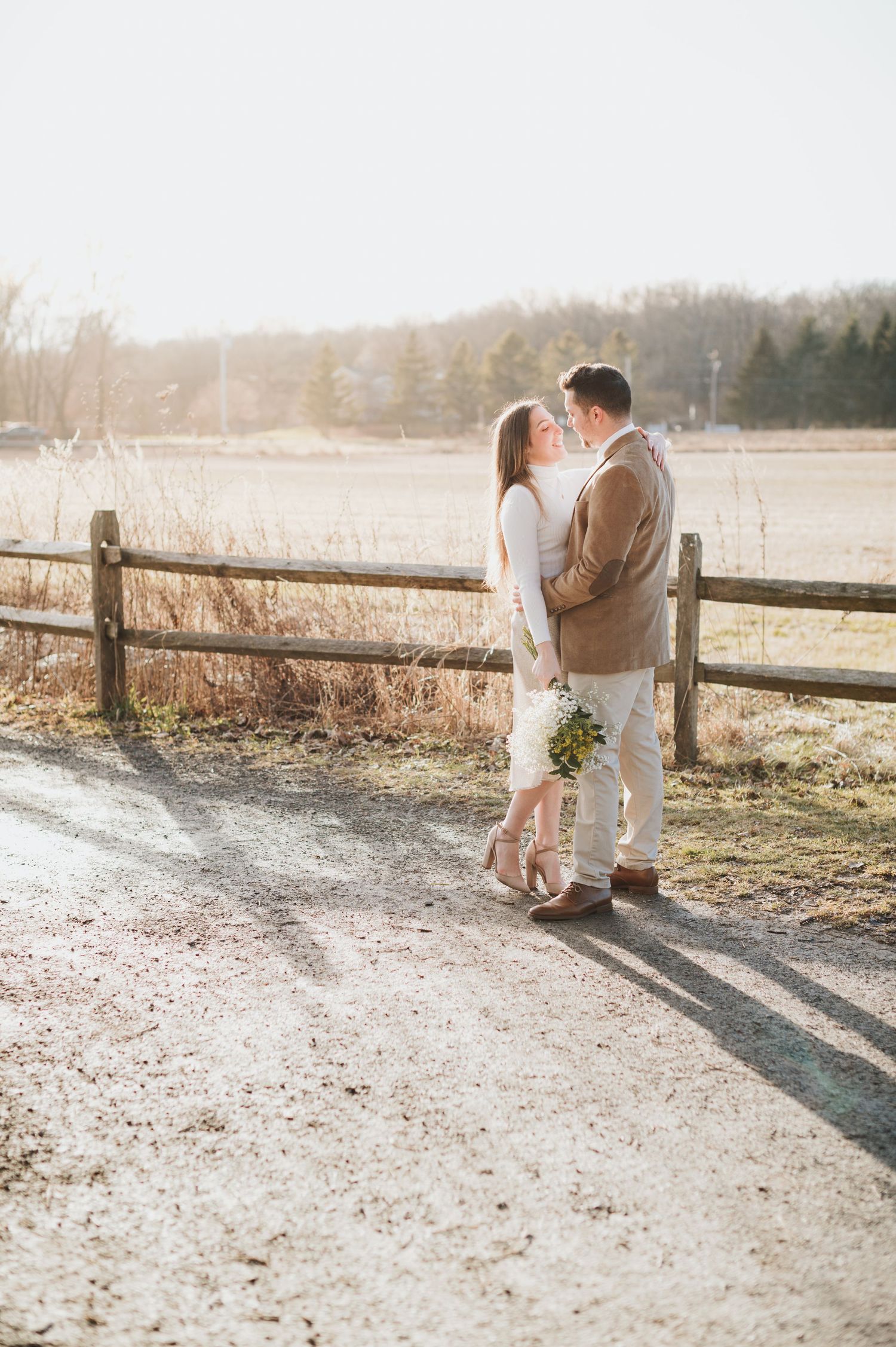 Couple shares intimate moment by wooden fence at sunset with delicate bouquet.