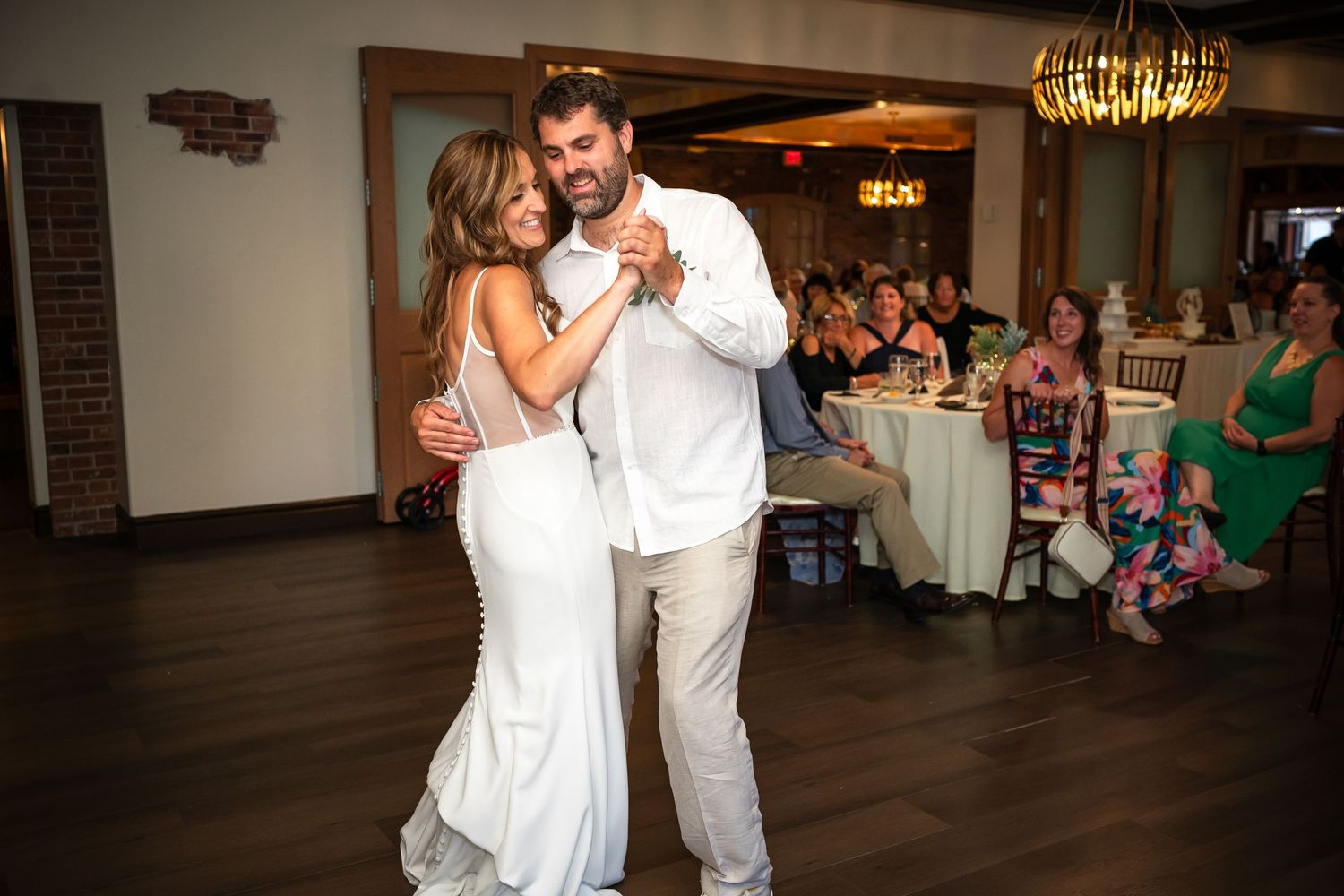 A couple shares a dance together at a wedding reception with guests seated at tables in the background.