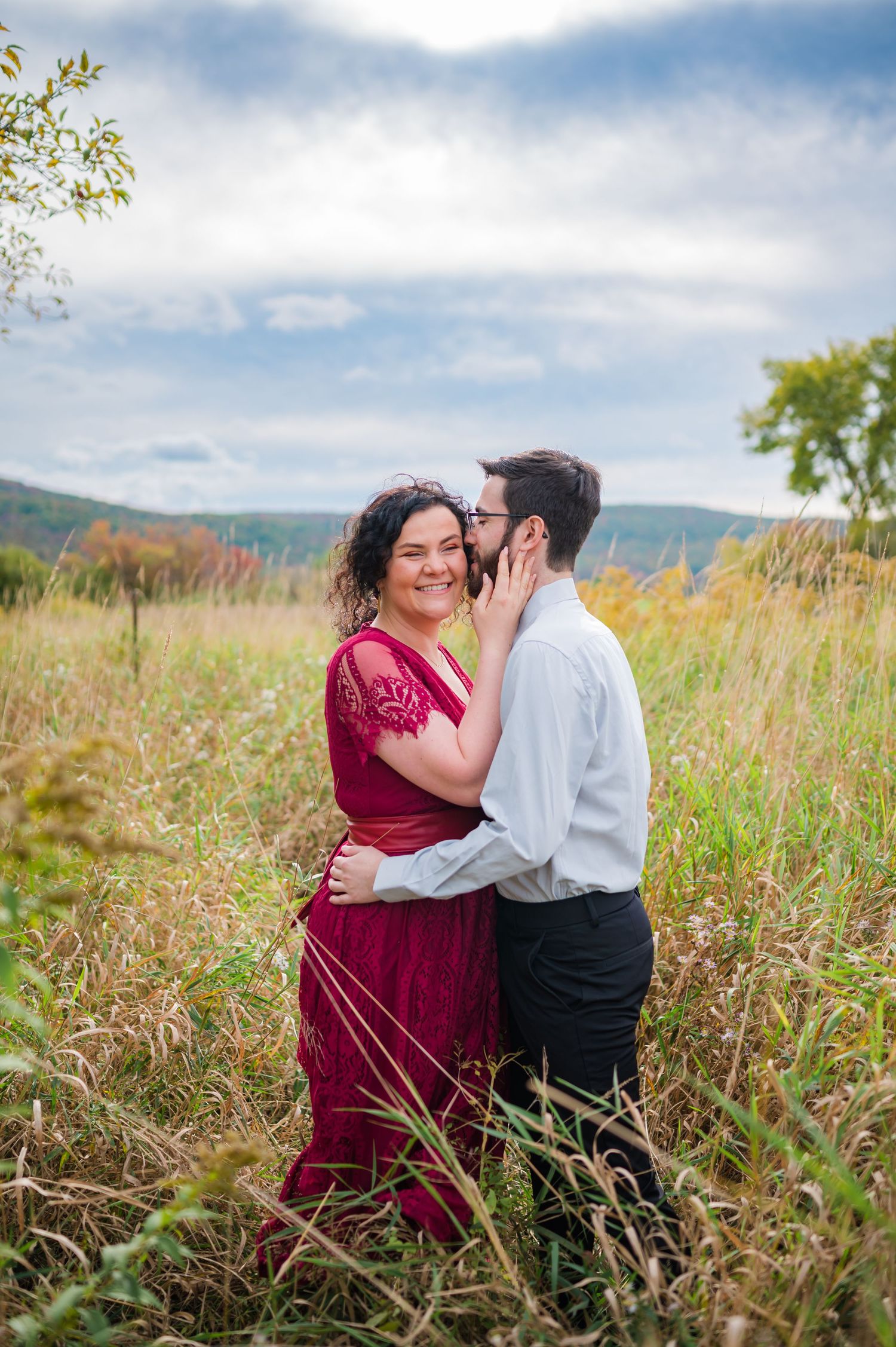 A couple embraces in a sunlit meadow with mountains in the background, she wearing a burgundy dress and he in white.