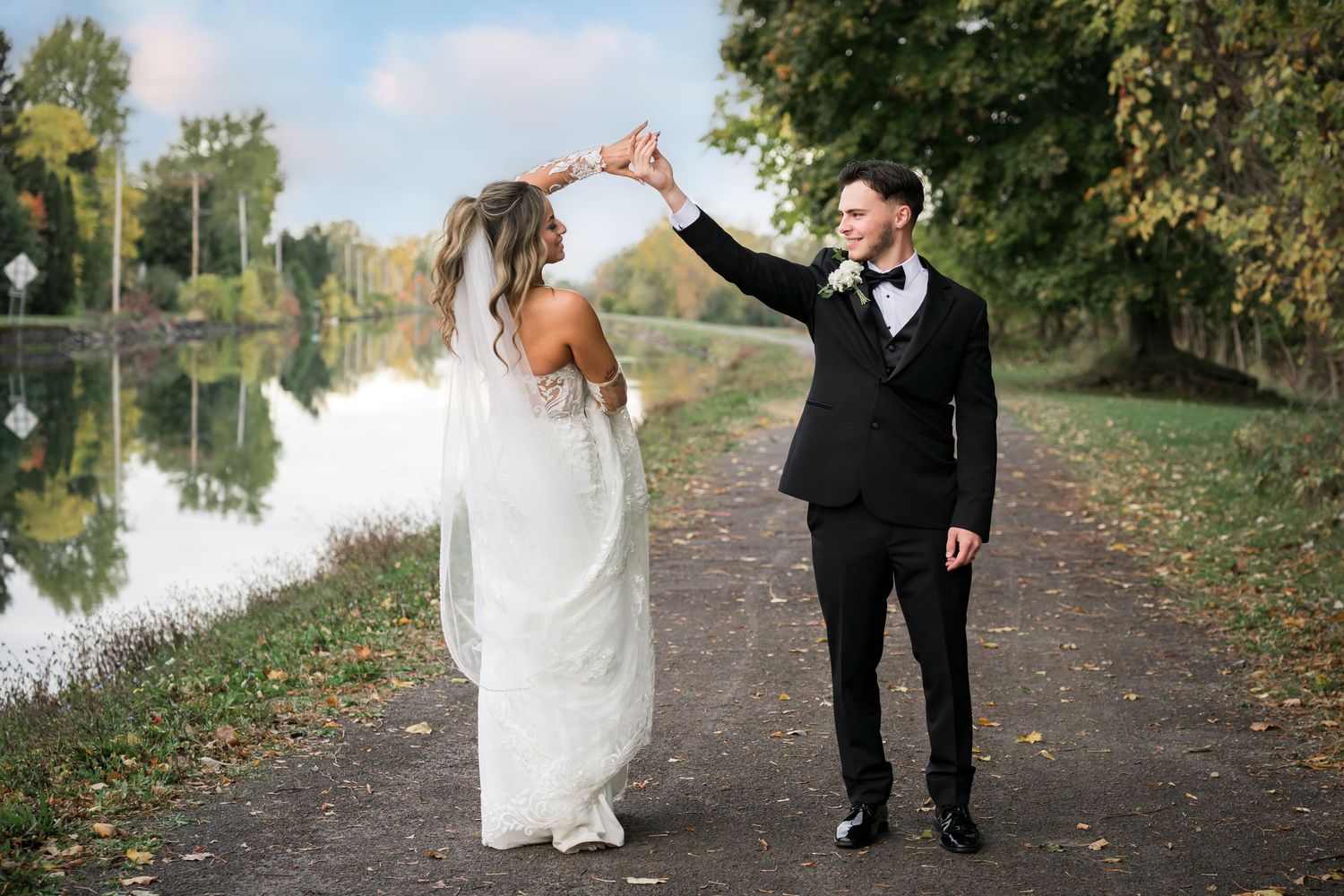 A couple in formal wedding attire walks along a tree-lined path by a lake on an autumn day.