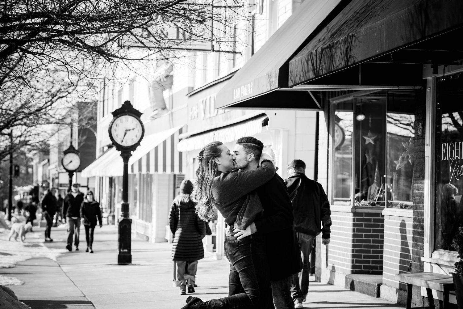 Couple kissing on a busy city sidewalk in black and white near a vintage street clock.