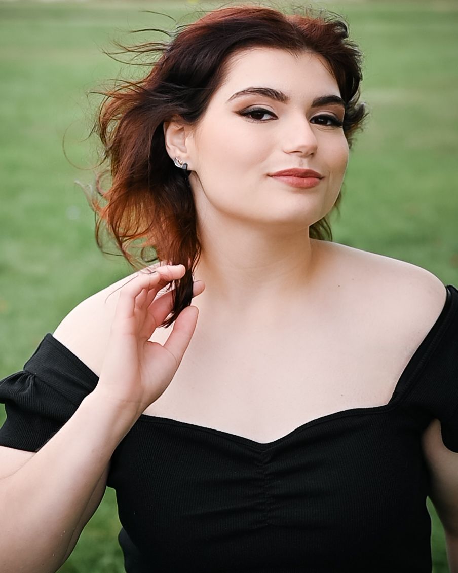 Outdoor portrait with windswept hair in black off-shoulder dress against green background.