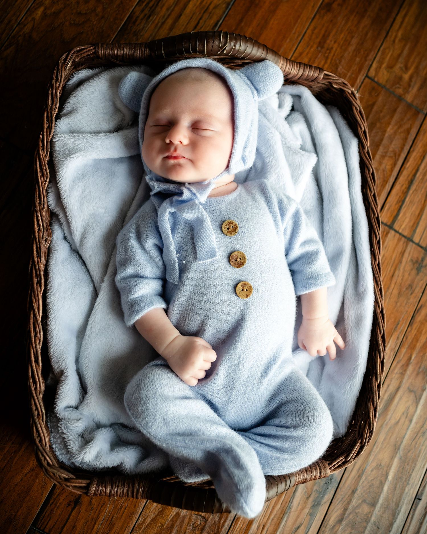 A sleeping newborn in a light blue knit romper with wooden buttons rests peacefully in a woven basket.