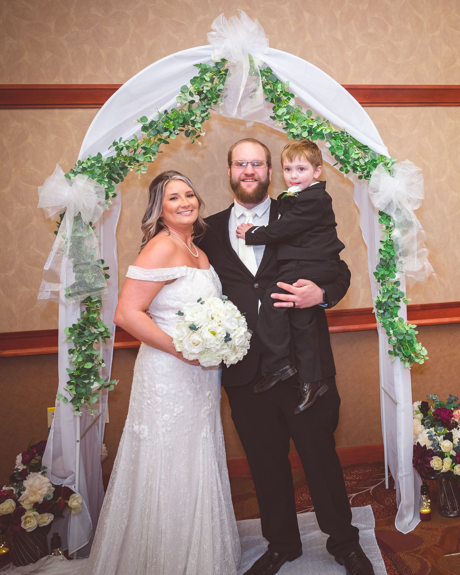 A wedding portrait under a decorated arch with tulle and greenery during an indoor ceremony.
