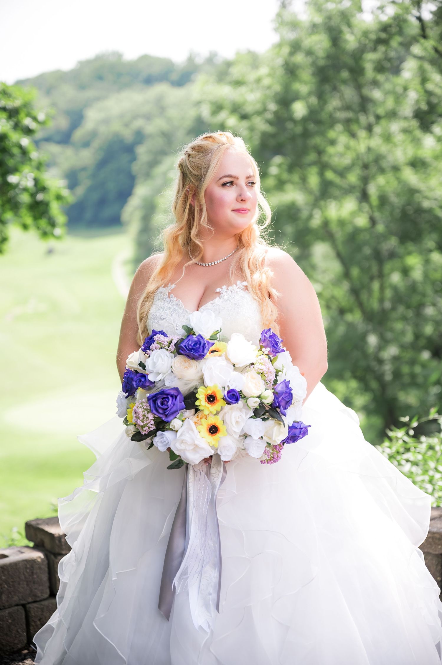 Wedding portrait featuring bride in tulle gown holding purple and sunflower bouquet against scenic backdrop.