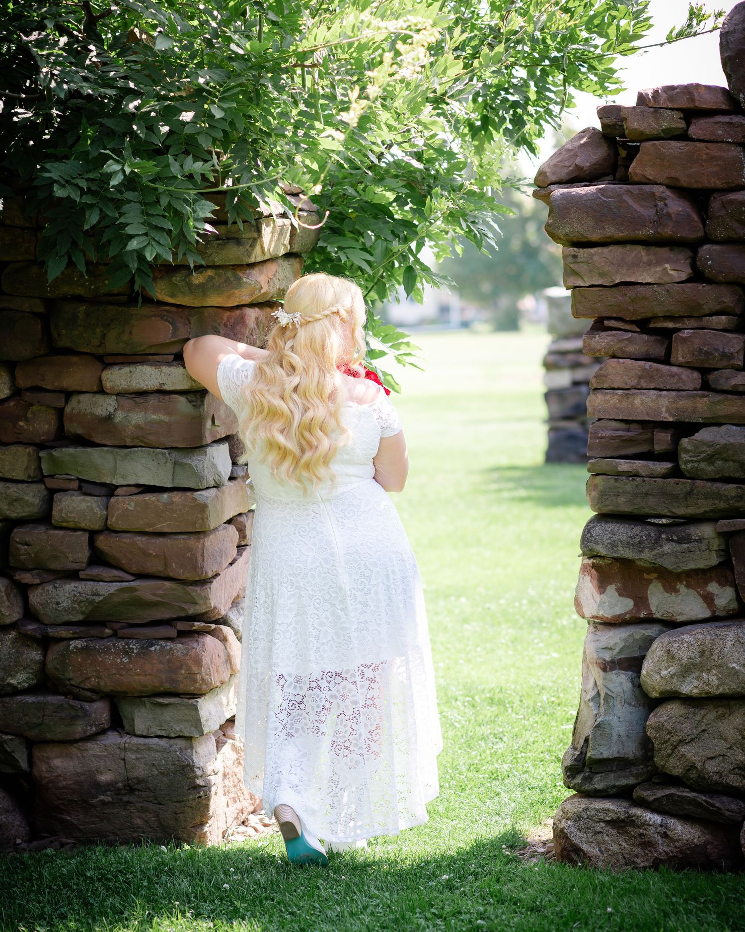 A white lace dress flows gracefully between stone archways in a sunlit garden setting.