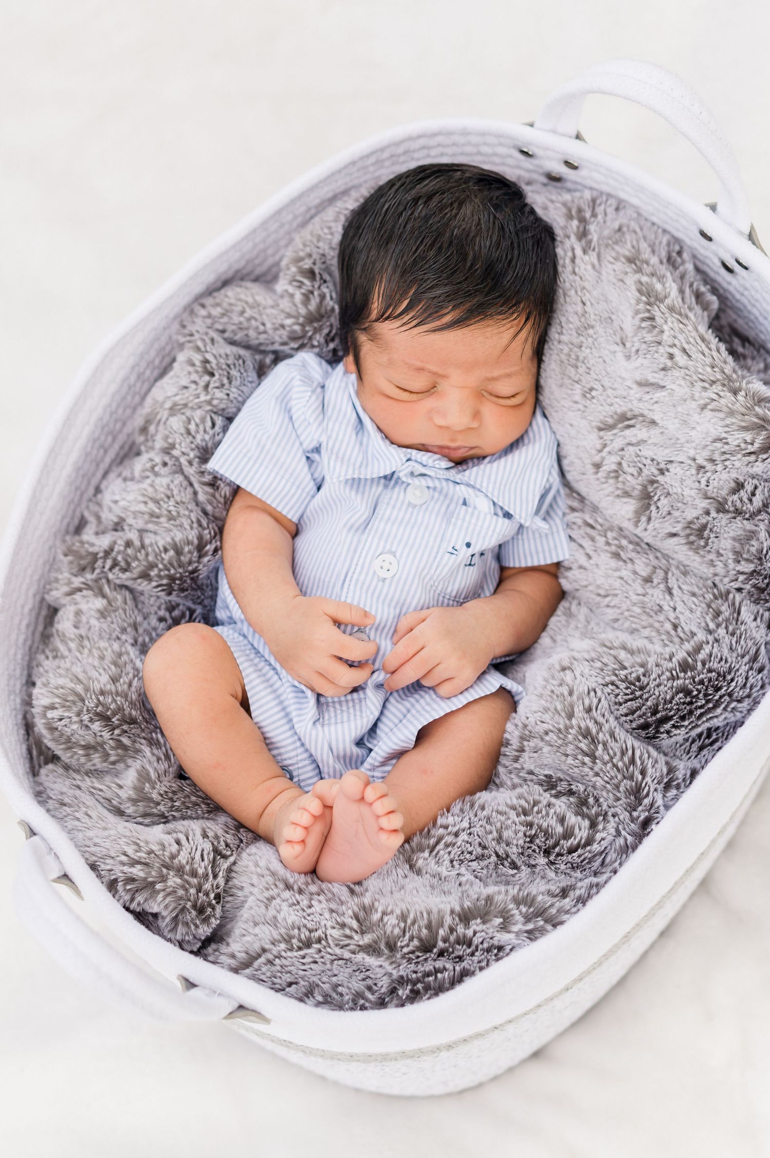 Sleeping newborn baby wearing blue striped outfit rests peacefully in white basket with plush gray blanket.