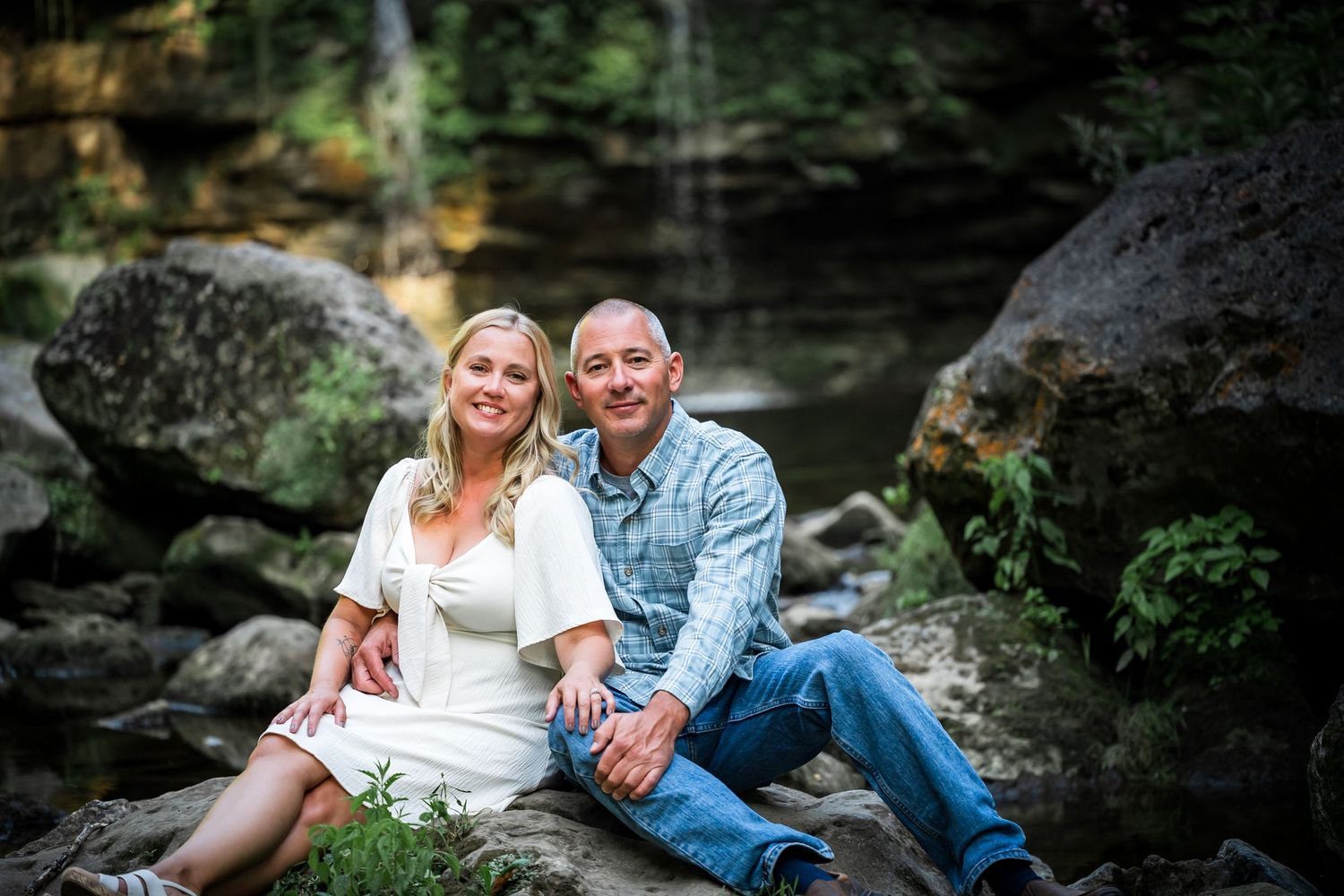 A couple sits together on rocks by a waterfall in a natural setting, wearing casual and white attire.