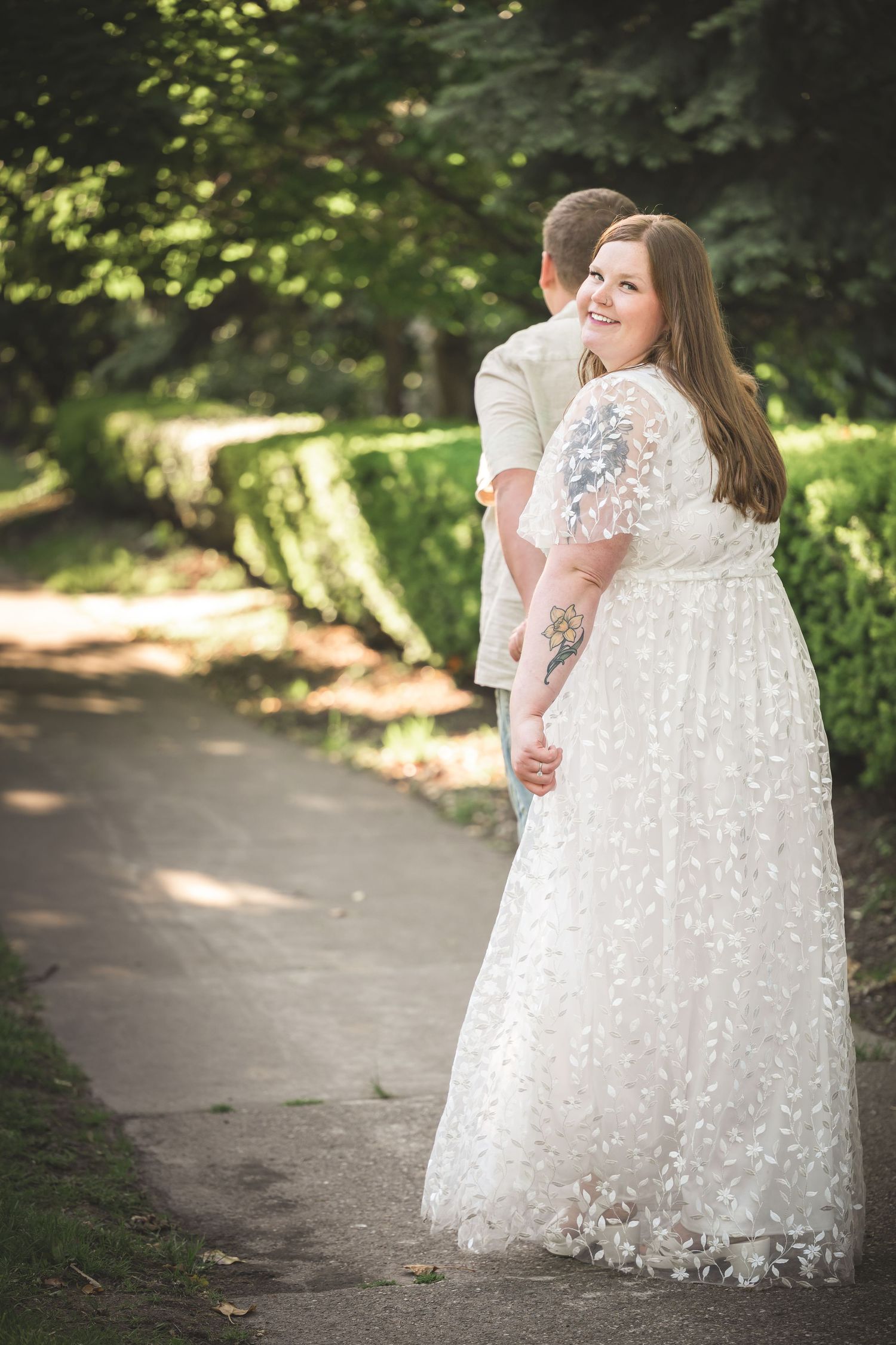 Bride in lace gown smiling over shoulder while groom embraces her on a garden path.