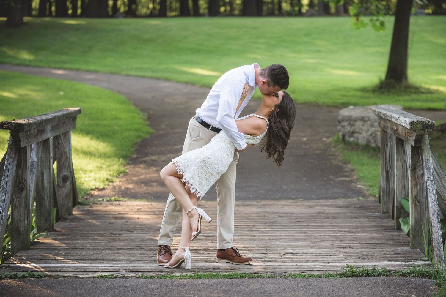 Couple sharing a romantic dip kiss on a wooden bridge in a lush green park during an engagement session.