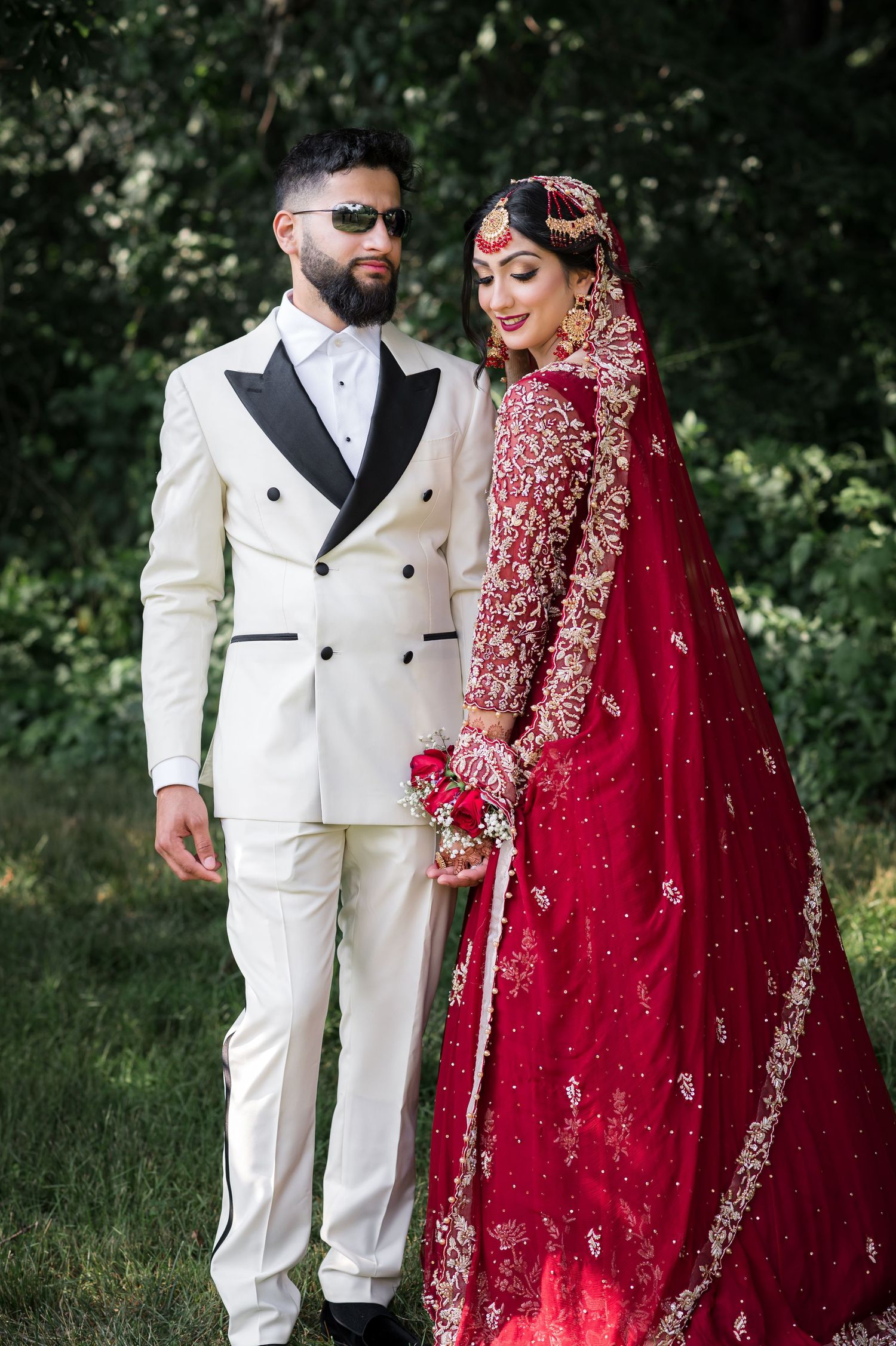 Wedding couple in white suit and red embroidered lehenga pose outdoors for romantic photos.