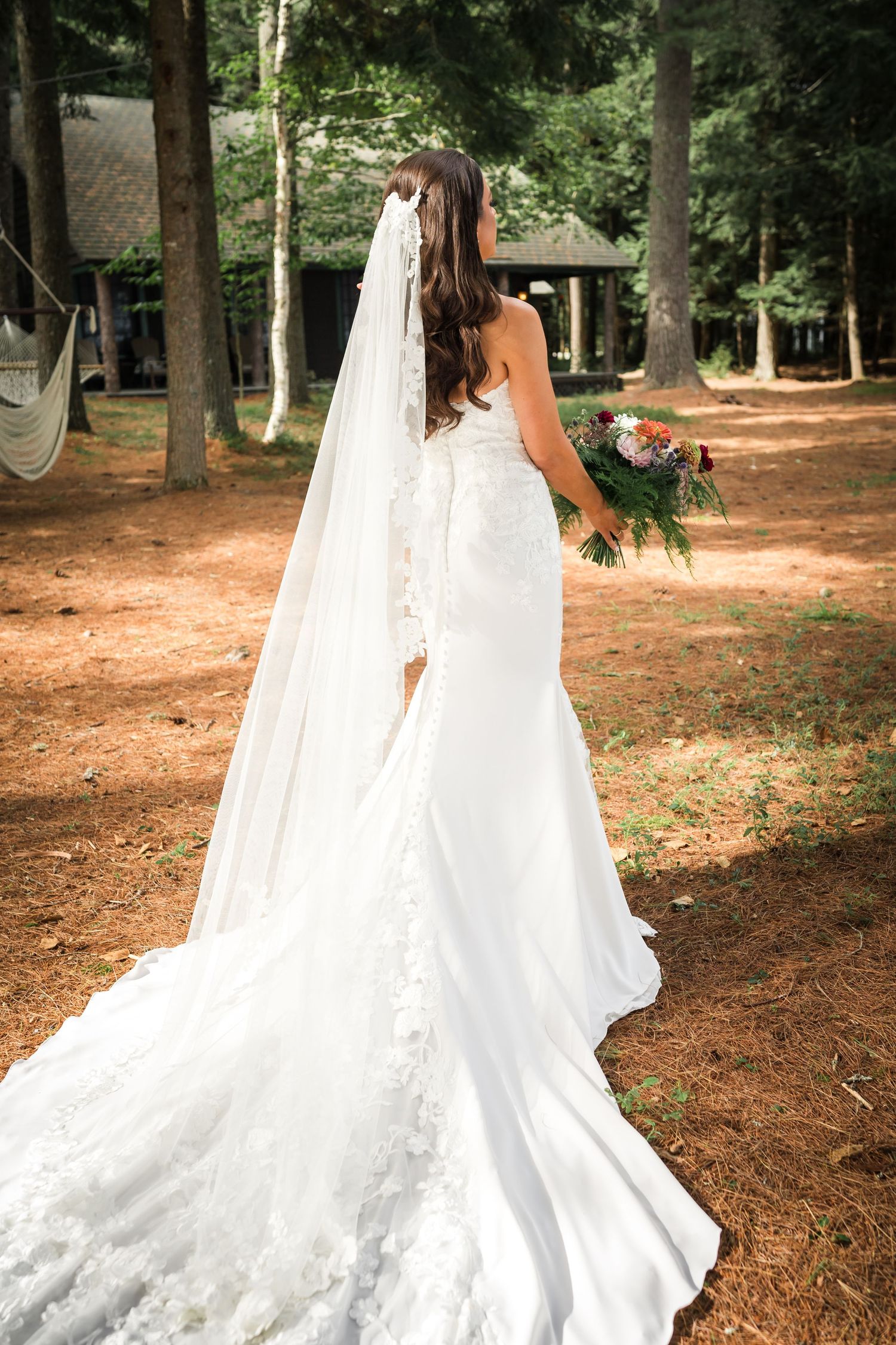 A bride in a flowing lace gown with cathedral veil walks through a wooded setting holding a bouquet.