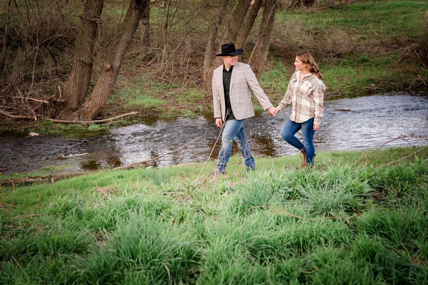 Couple walking hand in hand through green spring meadow beside flowing stream.