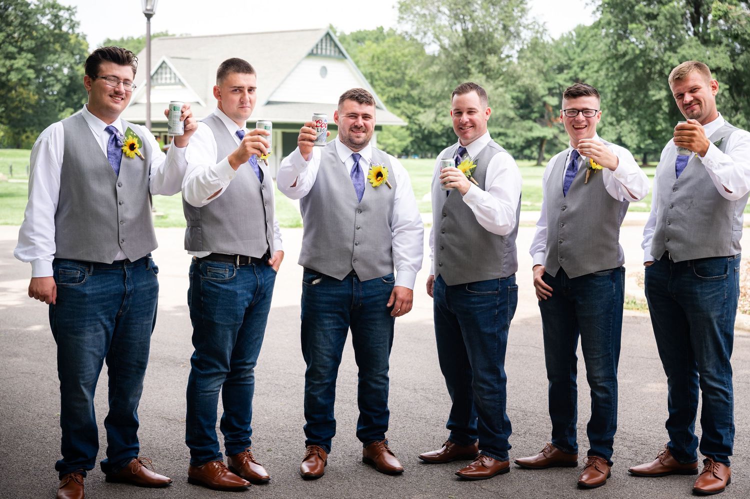 Groomsmen in matching gray vests and jeans pose while holding beverages.