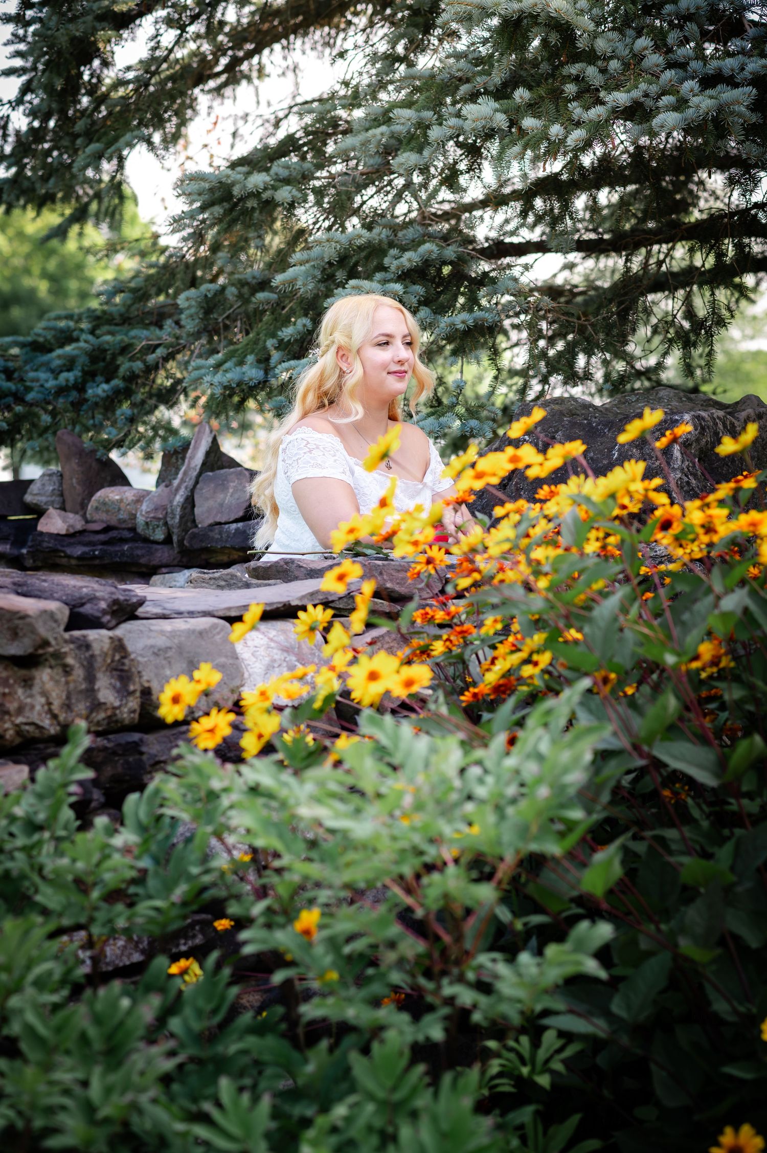 A white-dressed figure sits among vibrant yellow flowers and evergreen trees in a garden setting.
