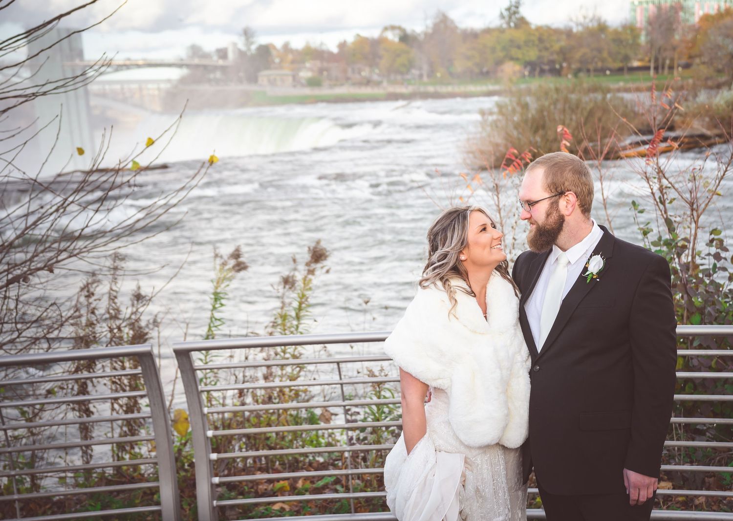 Wedding couple shares intimate moment on riverside deck with fall foliage and water views.