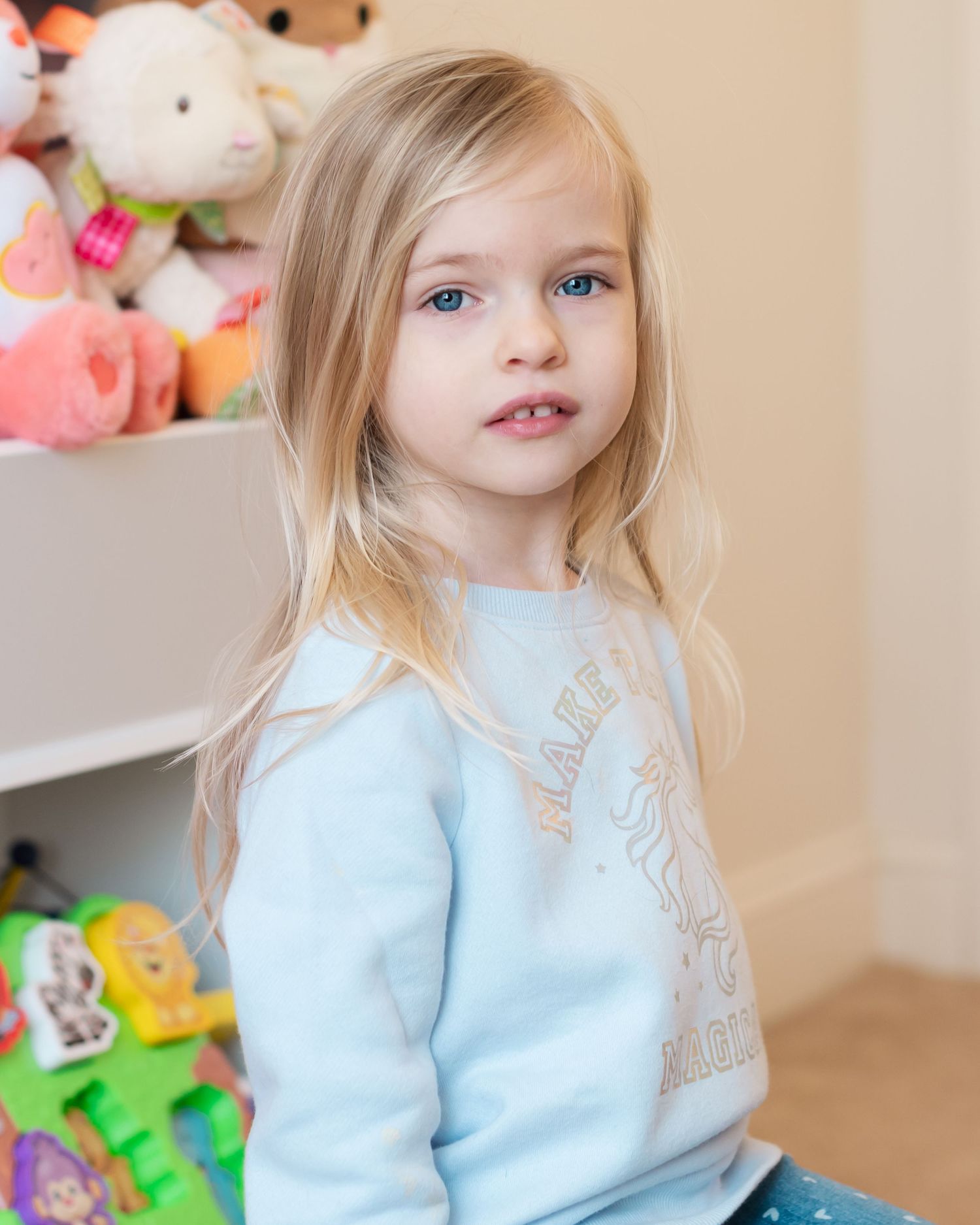 Child in a light blue sweater posing near toy shelf with stuffed animals.