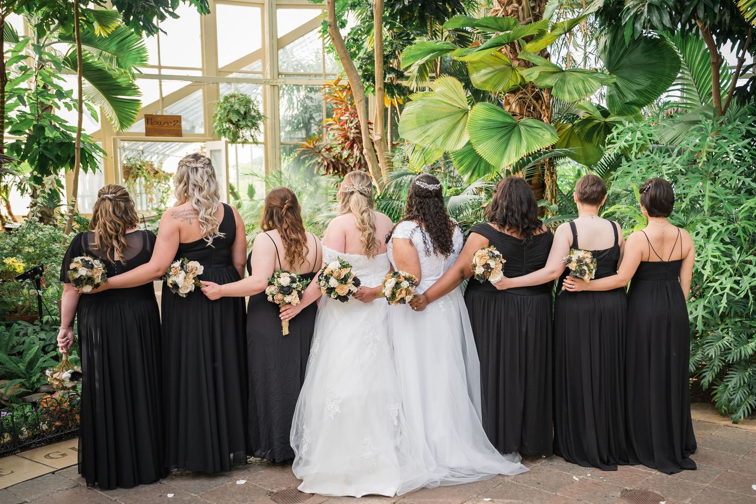 Wedding party in black dresses and white gowns pose together in a greenhouse setting.