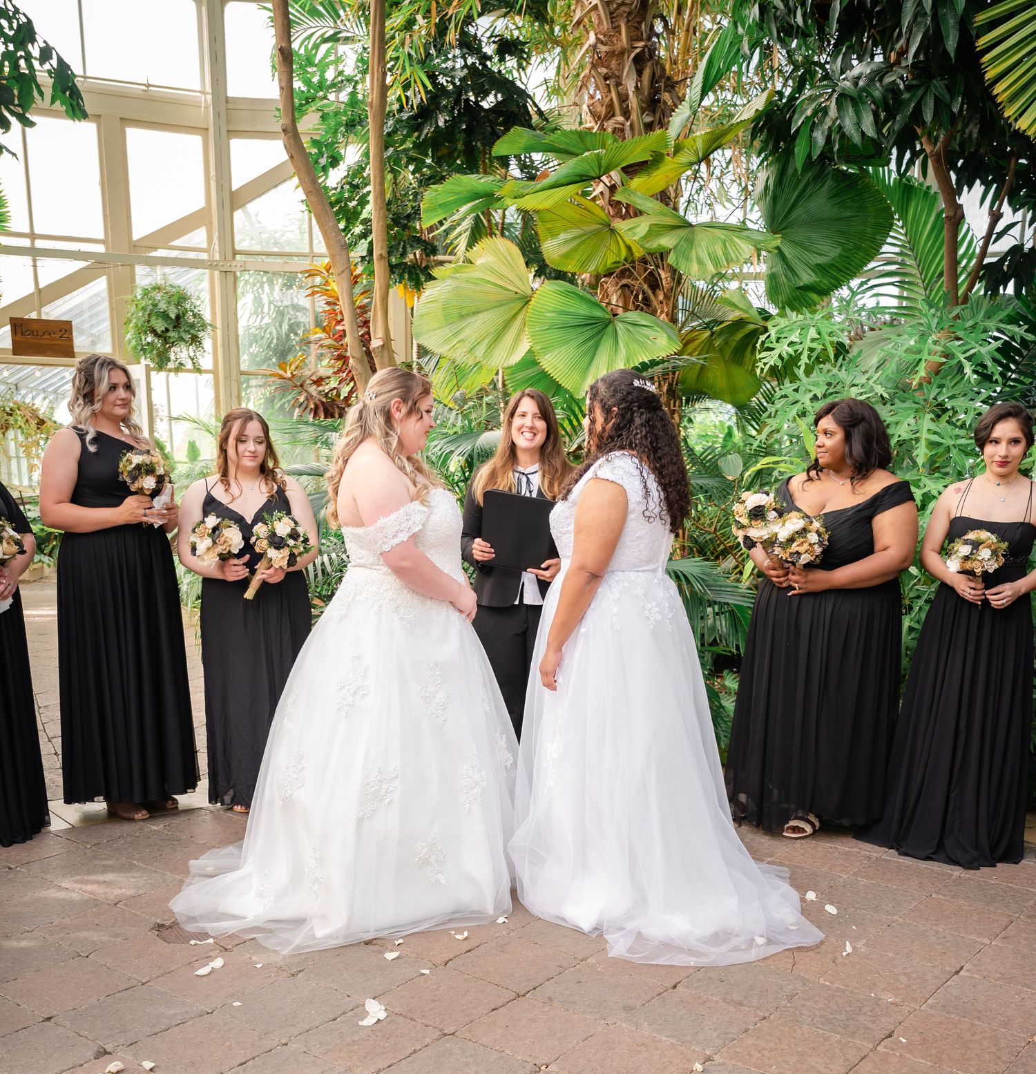Wedding party in black dresses surrounds couple in white gowns during greenhouse ceremony.
