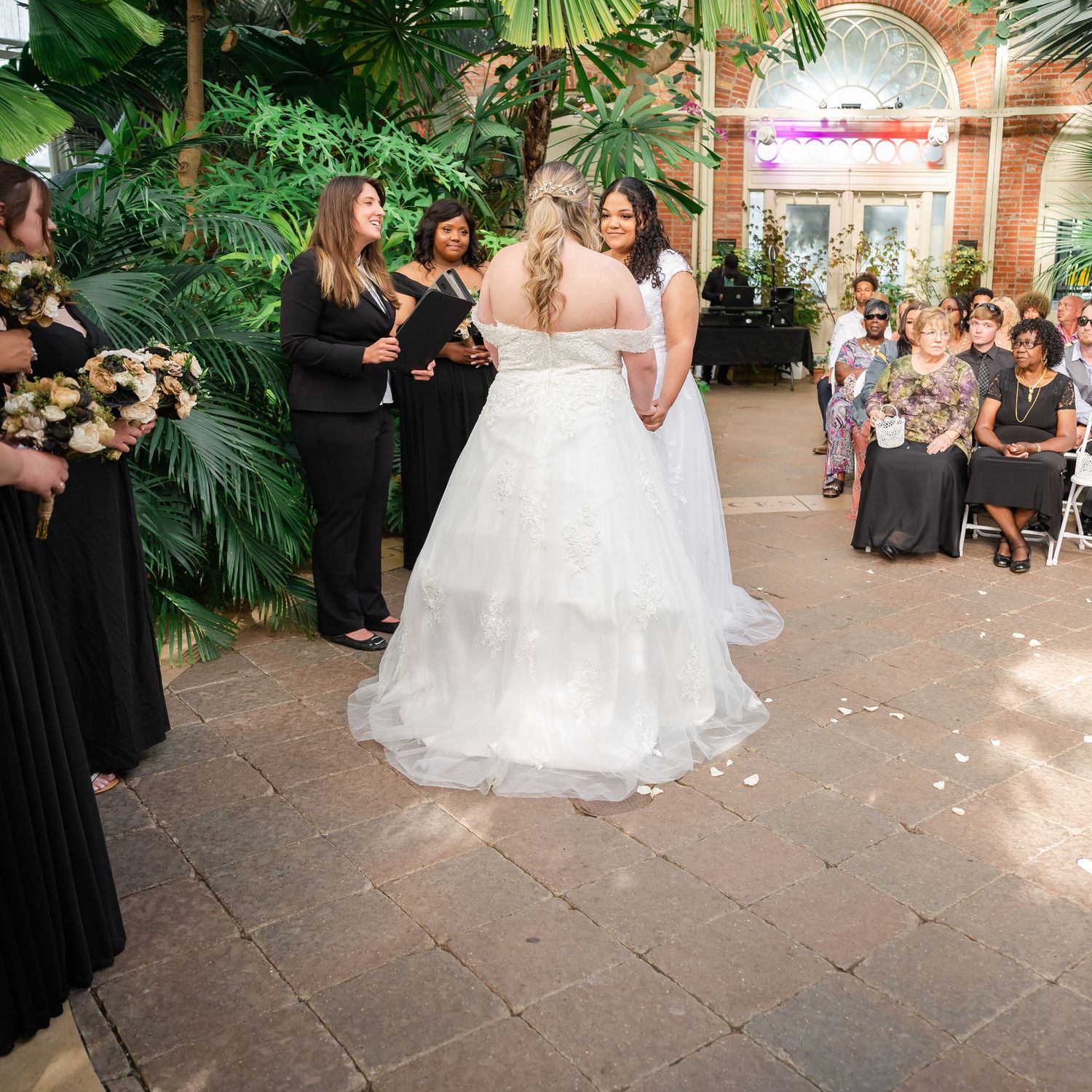 Wedding ceremony in a lush conservatory with attendees in black dresses watching the couple exchange vows.