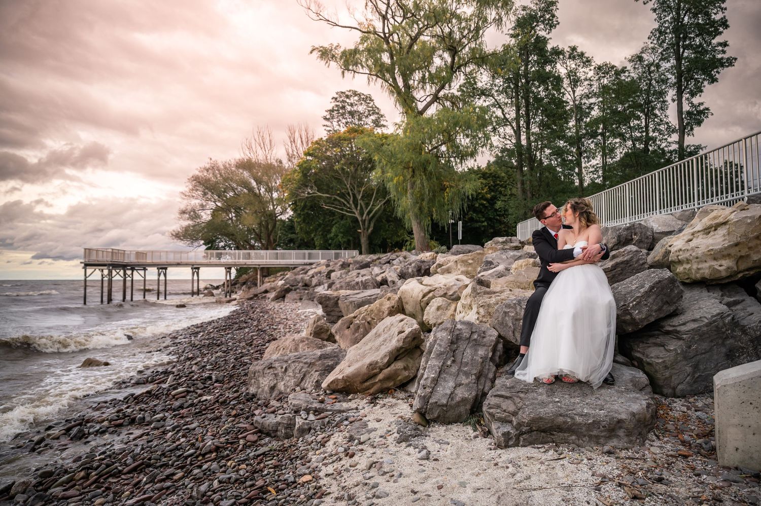 Wedding couple shares intimate moment on rocky beach with pier and dramatic sunset in background.