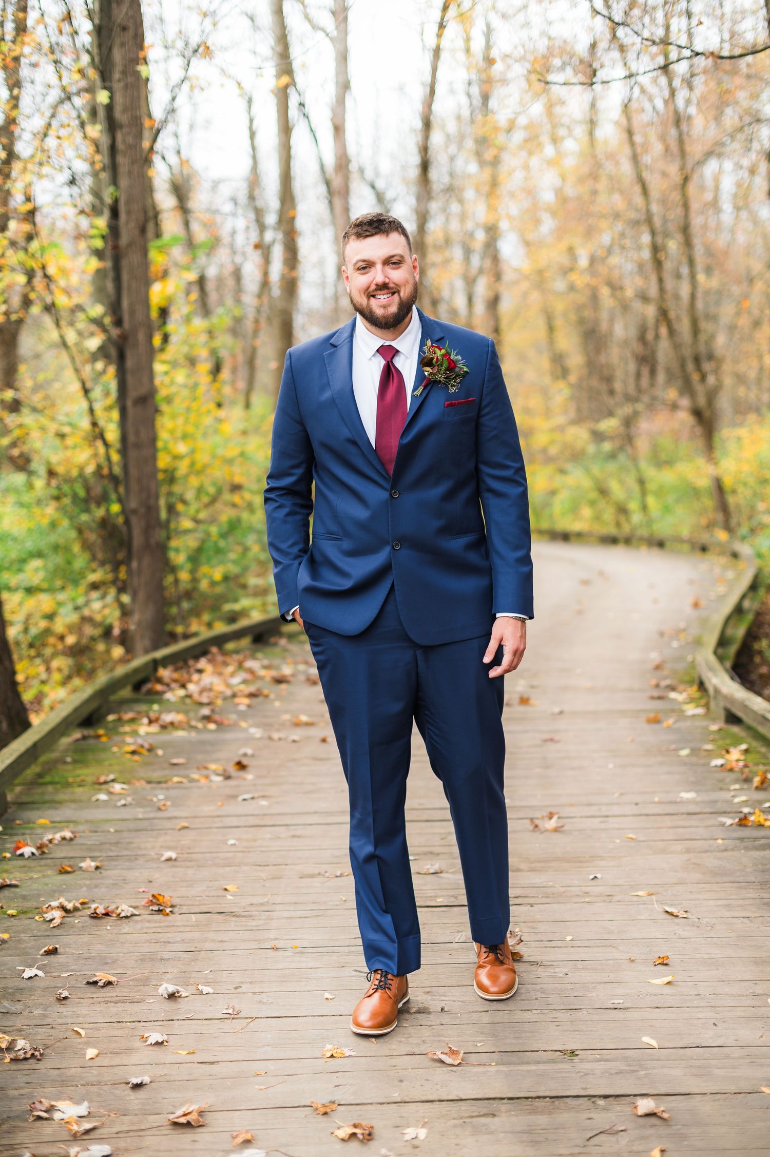 Groom in navy suit and red tie poses on wooden boardwalk during autumn.
