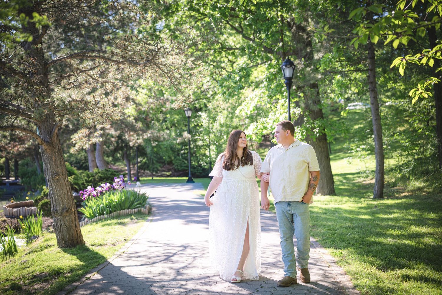 Couple walking hand in hand on a tree-lined park path, woman in white maternity dress, man in jeans on sunny day.