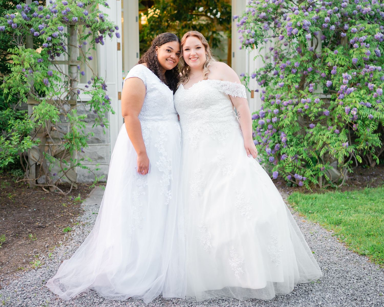 Two brides in beautiful white wedding gowns pose together in a garden setting with purple flowering vines.