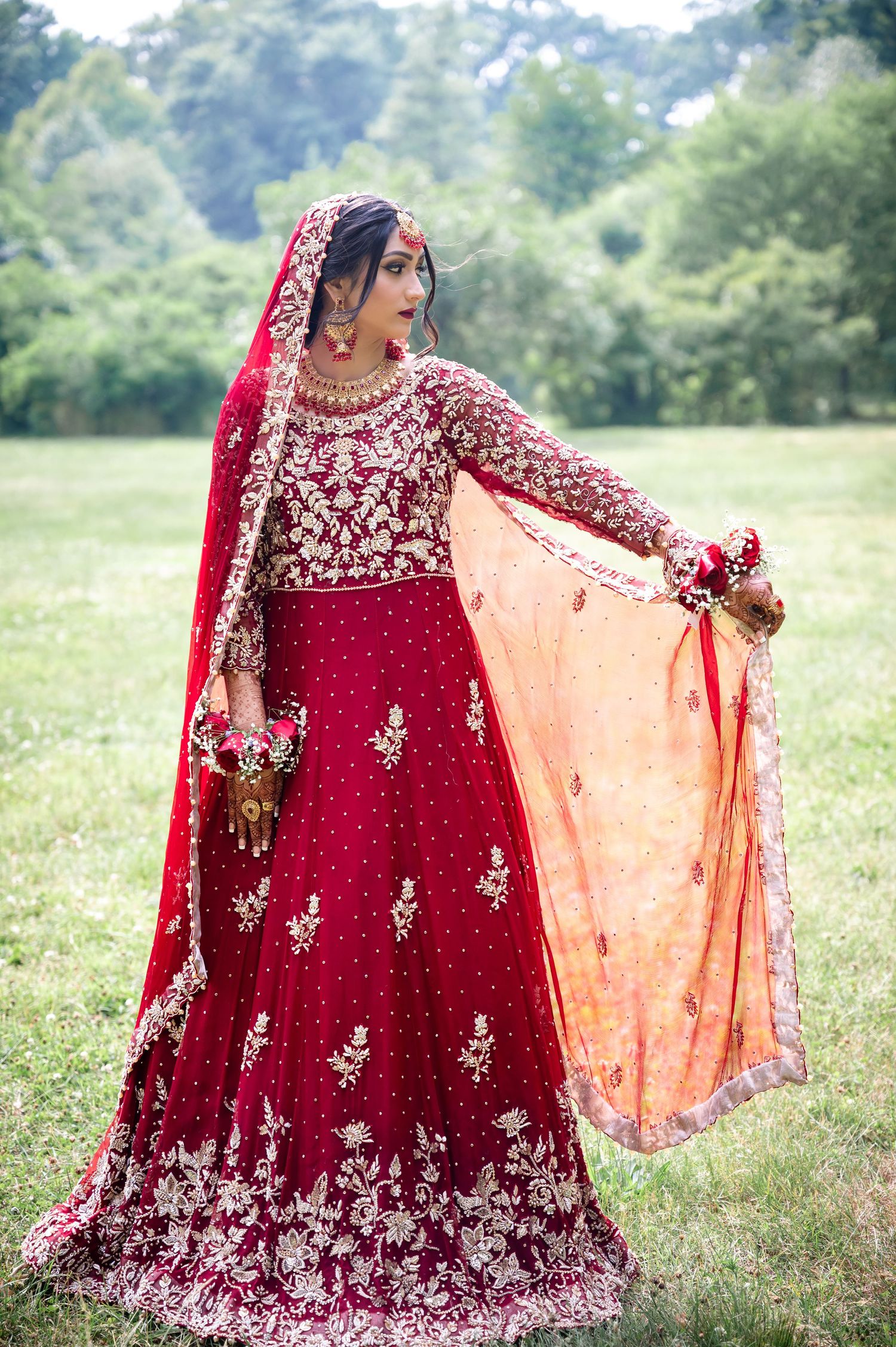 Traditional South Asian bride in ornate red lehenga with gold embroidery posing in garden setting.