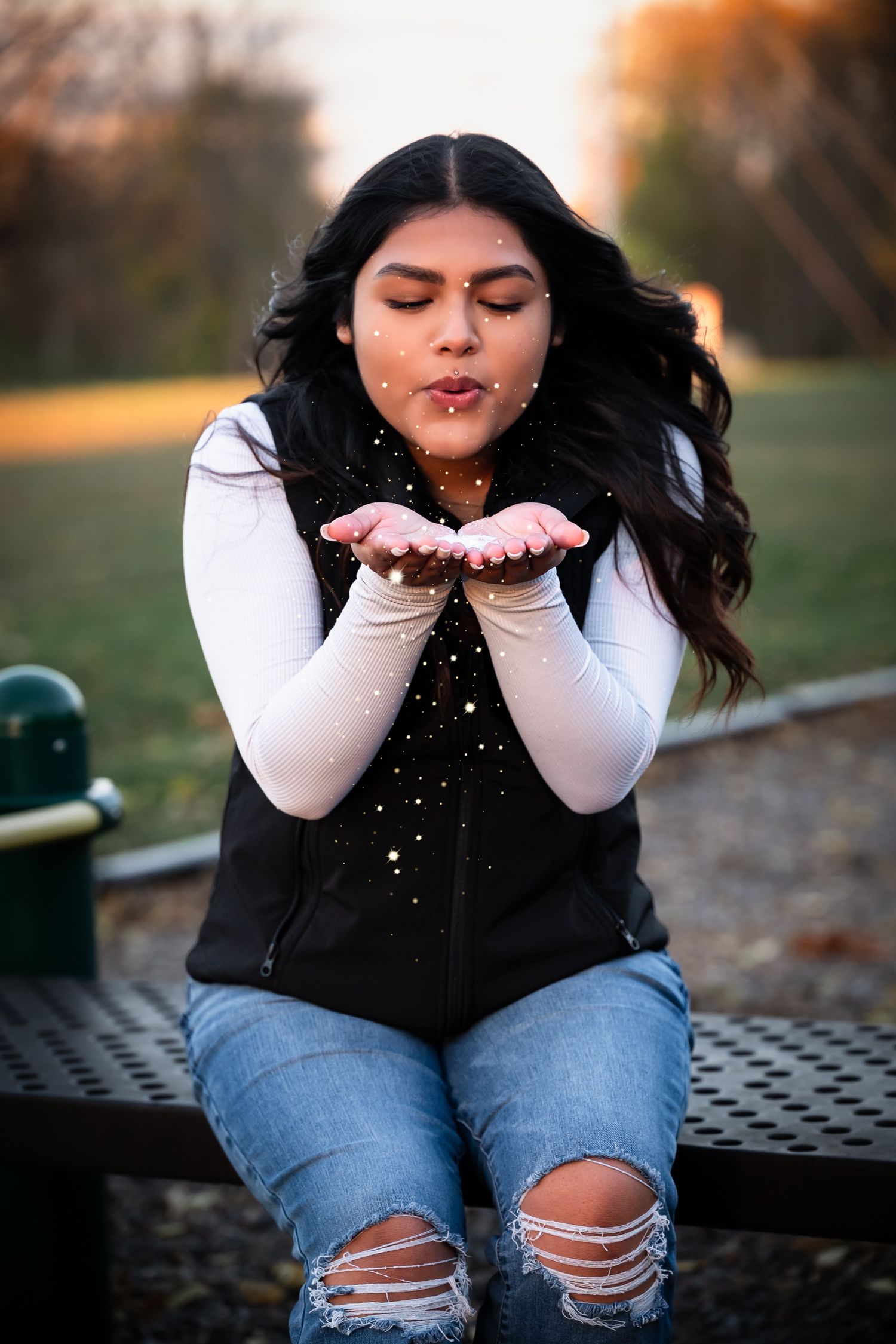 Person wearing ripped jeans and black vest blows glitter from their hands in an autumn outdoor photoshoot sequence.