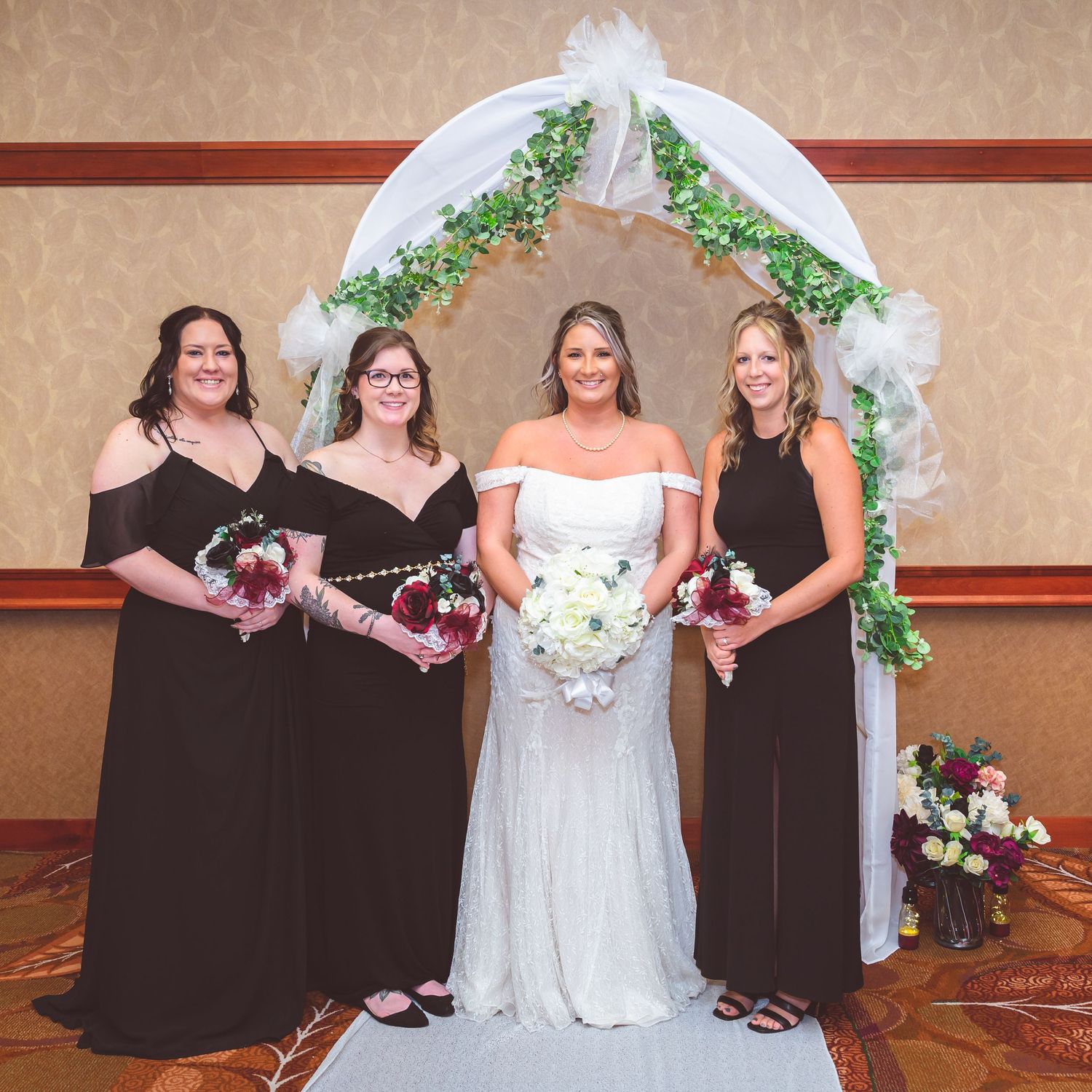 Four women in formal attire pose under a white wedding arch decorated with greenery and flowers.