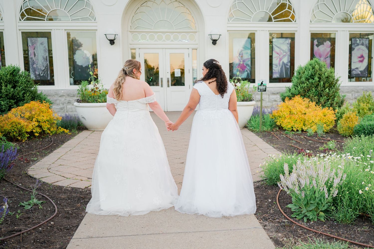 Two brides in white dresses hold hands while walking along a garden path.