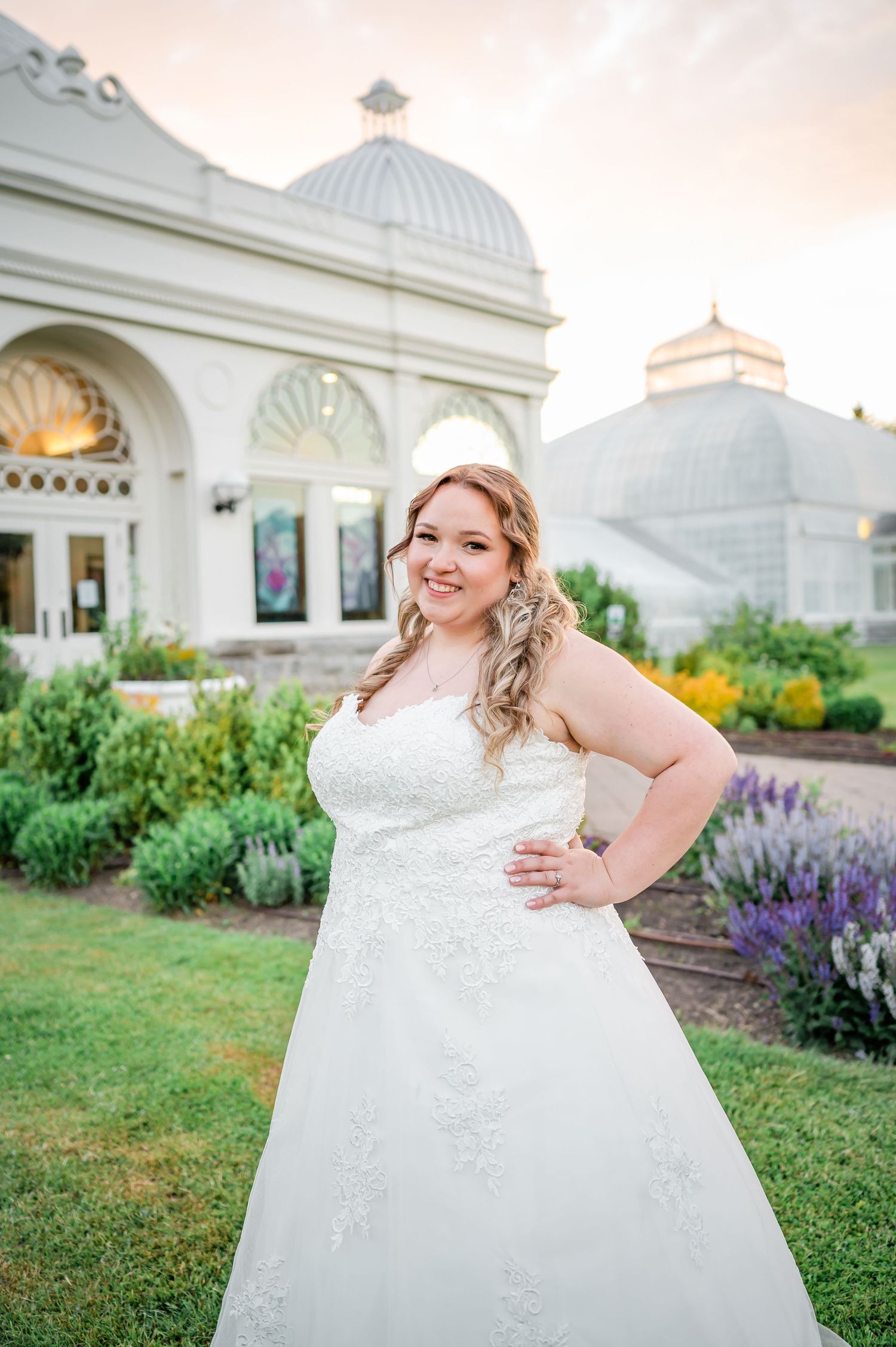 A bride in a lace wedding gown poses in front of a domed conservatory building with lavender garden.