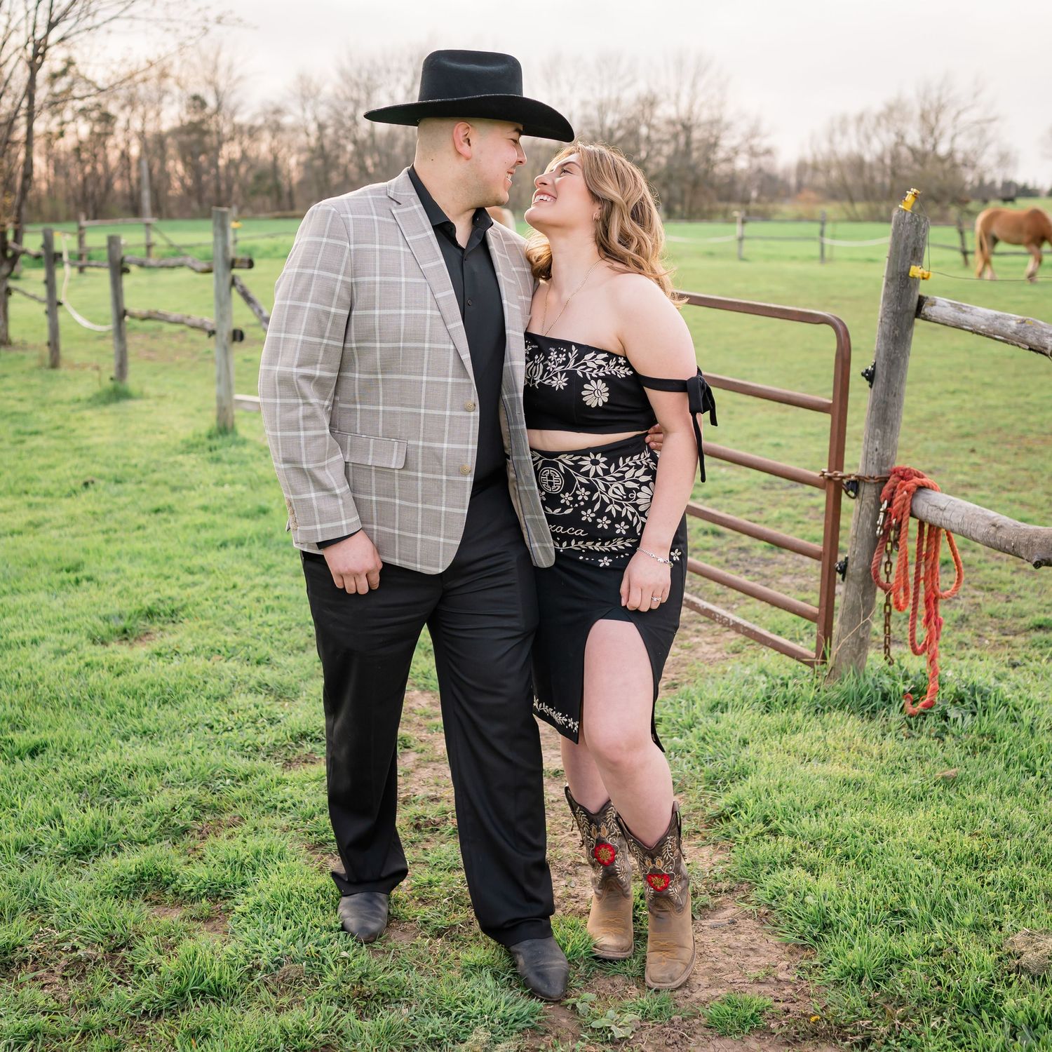 A couple poses romantically by a rustic fence in a pasture with horses visible in the background.