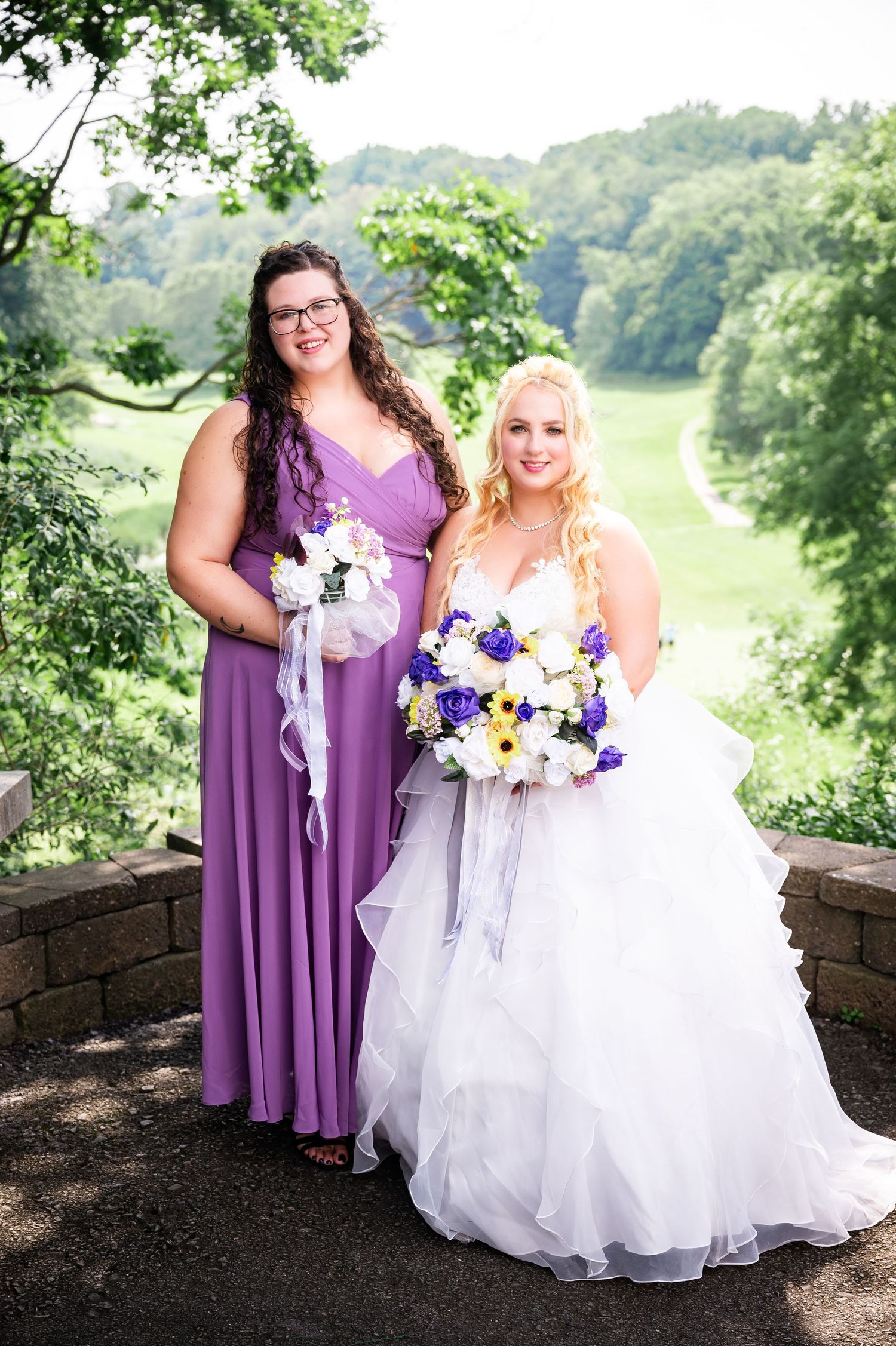 A bridal party member in purple and the bride posing with floral bouquets on a stone overlook.