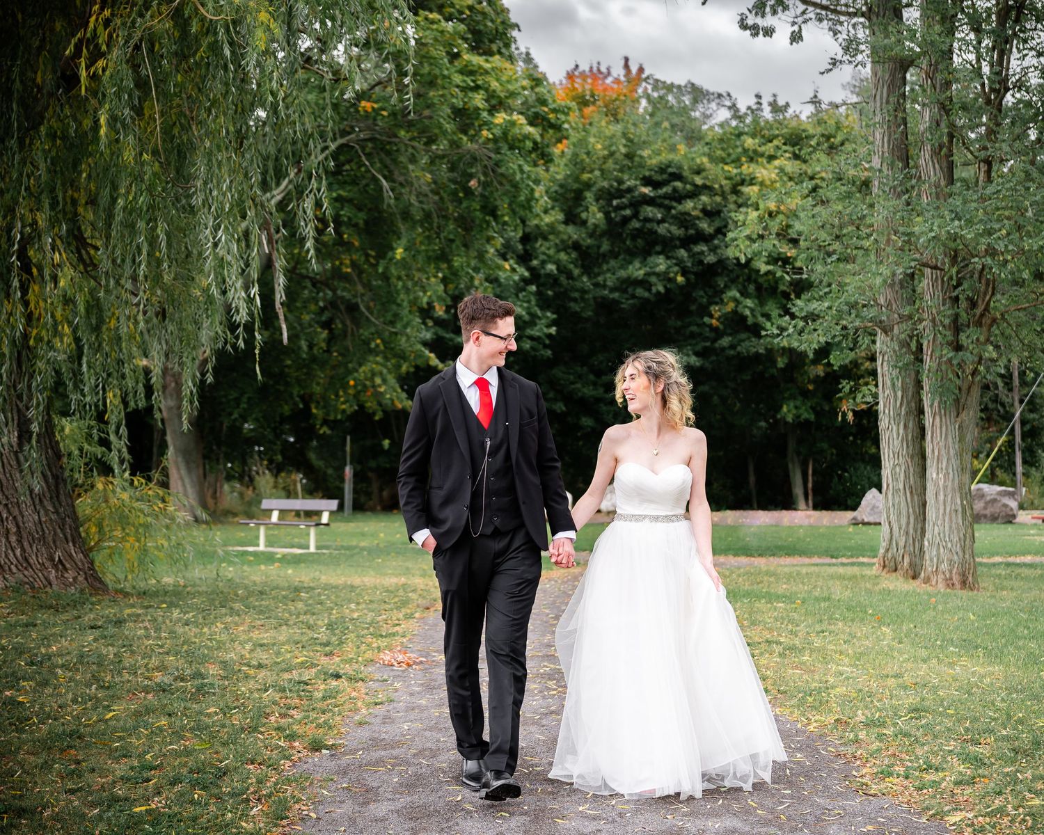 A couple walks hand in hand along a park path surrounded by willow trees and autumn foliage.