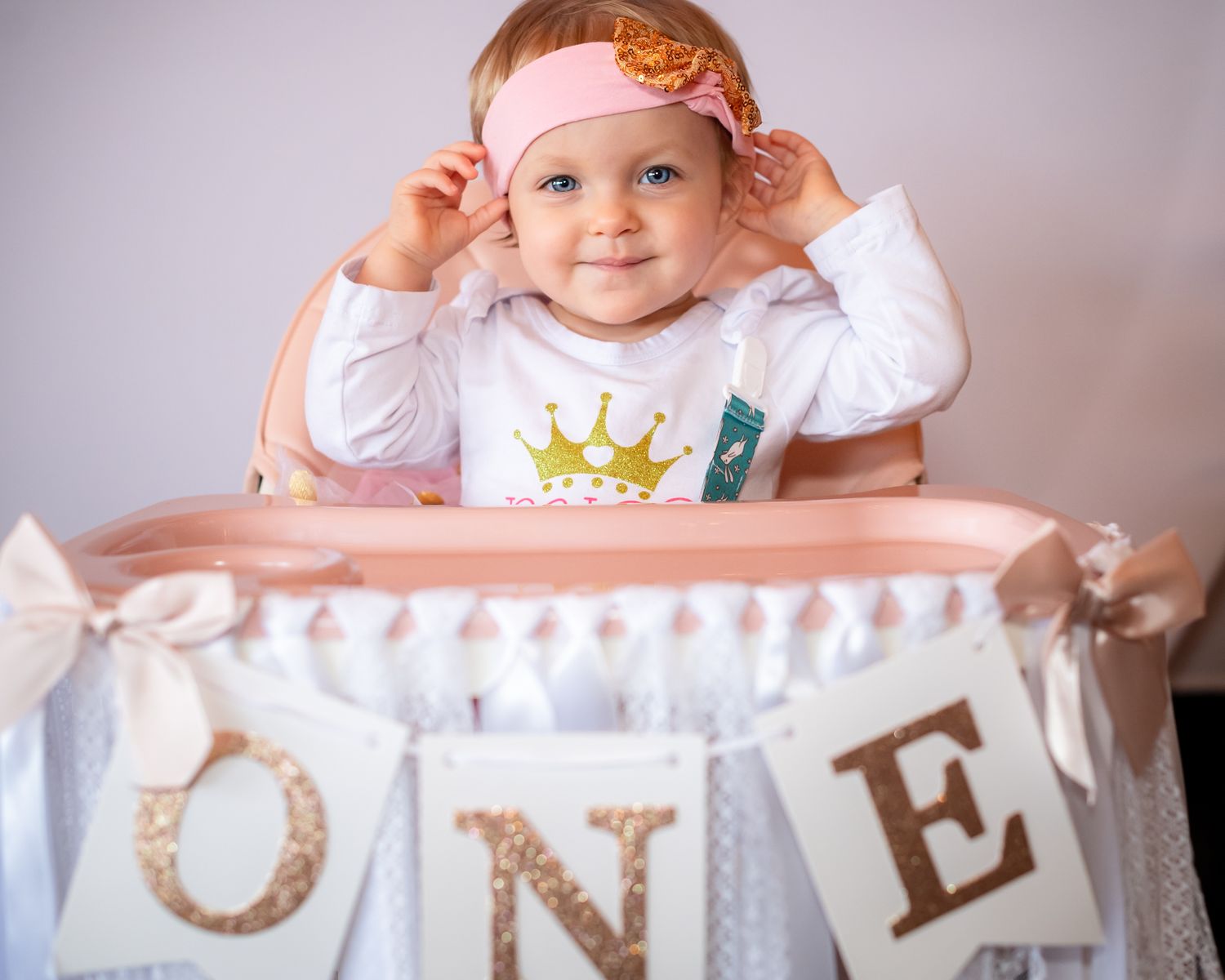 Baby wearing orange bow sits in high chair with 'ONE' banner decoration for first birthday celebration.