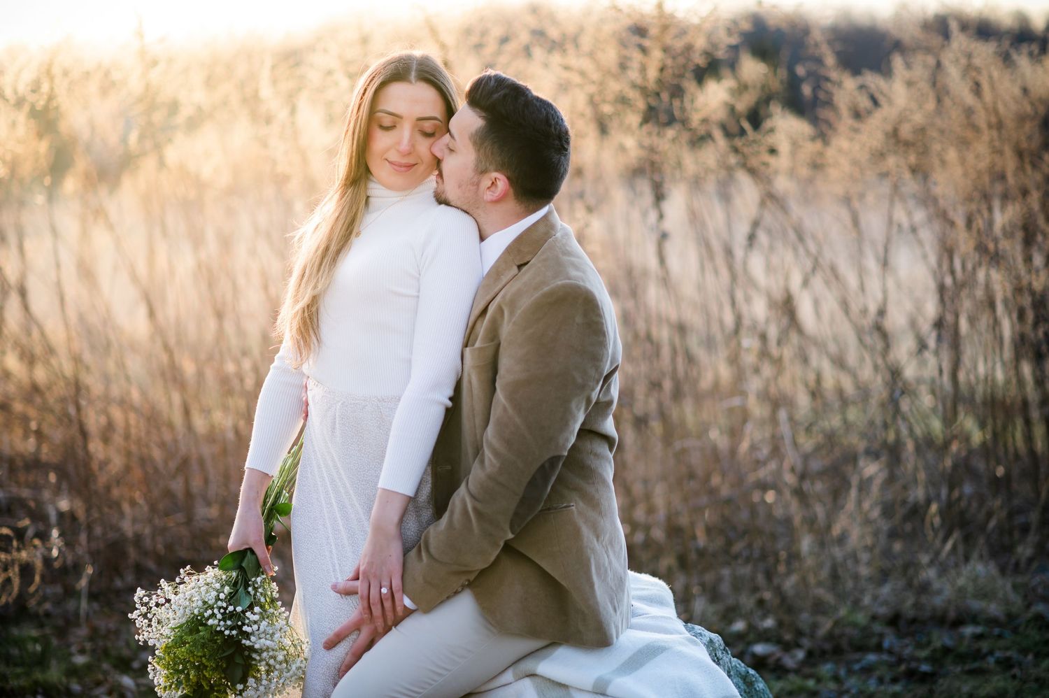 A couple in wedding attire shares an intimate moment in a golden field during sunset while holding a wildflower bouquet.