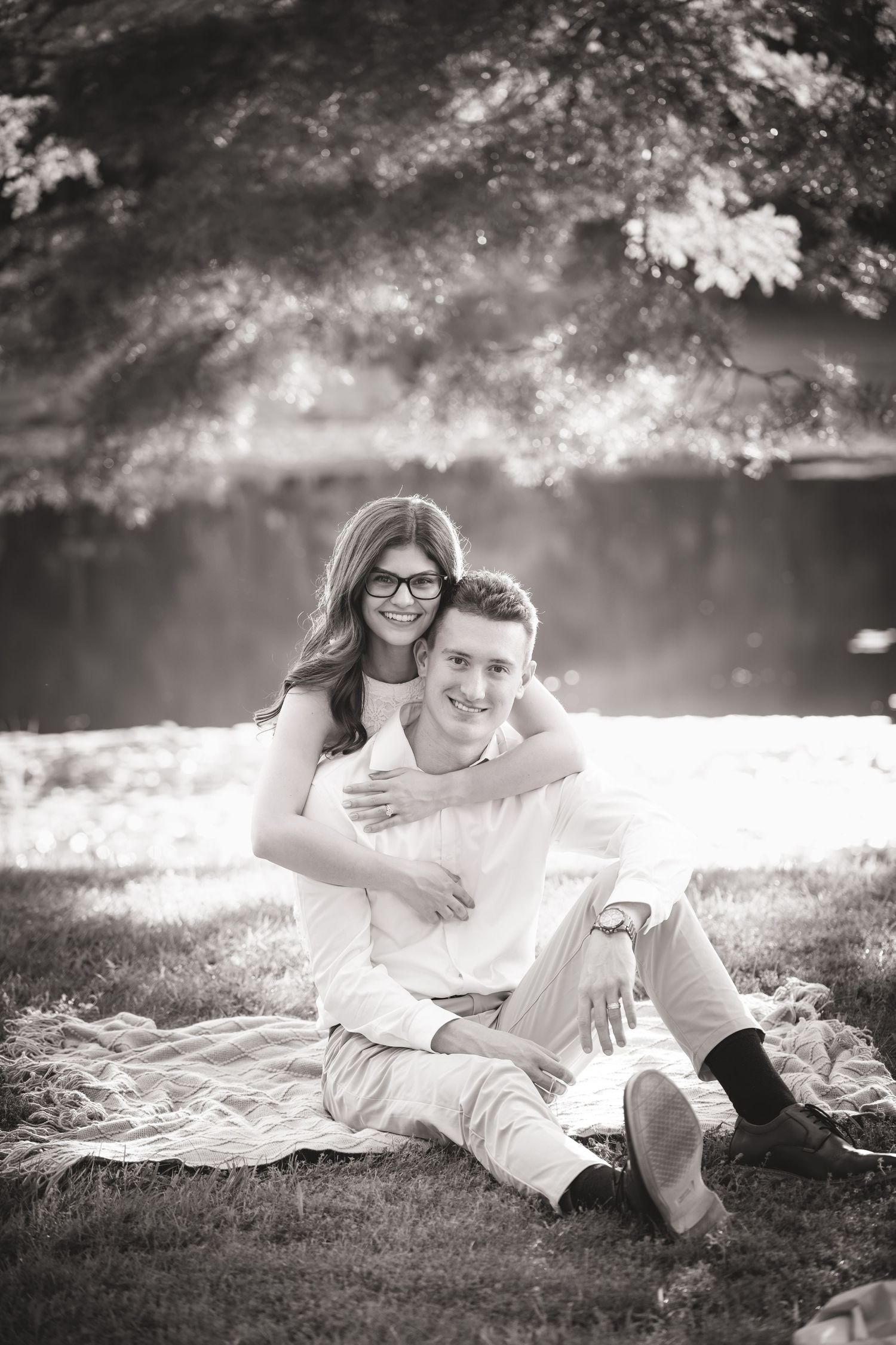 Black and white photo of a smiling couple sitting on a blanket outdoors, woman hugging man from behind near a sunlit lake.