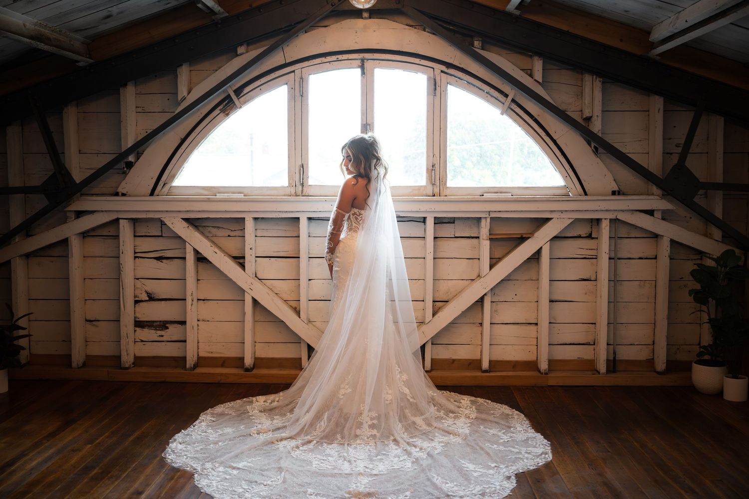 A bride in a flowing lace wedding gown and veil poses in a rustic wooden barn with natural light streaming through windows.