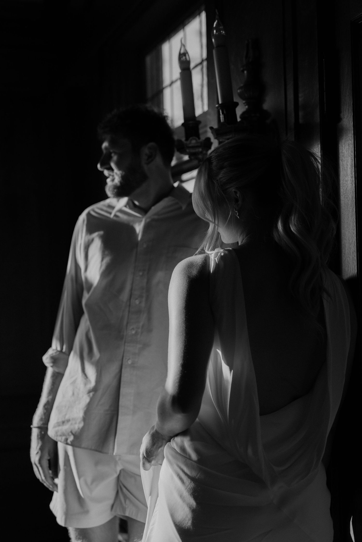 black and white photo couple leaning against a  window with moody lighting shining on them.