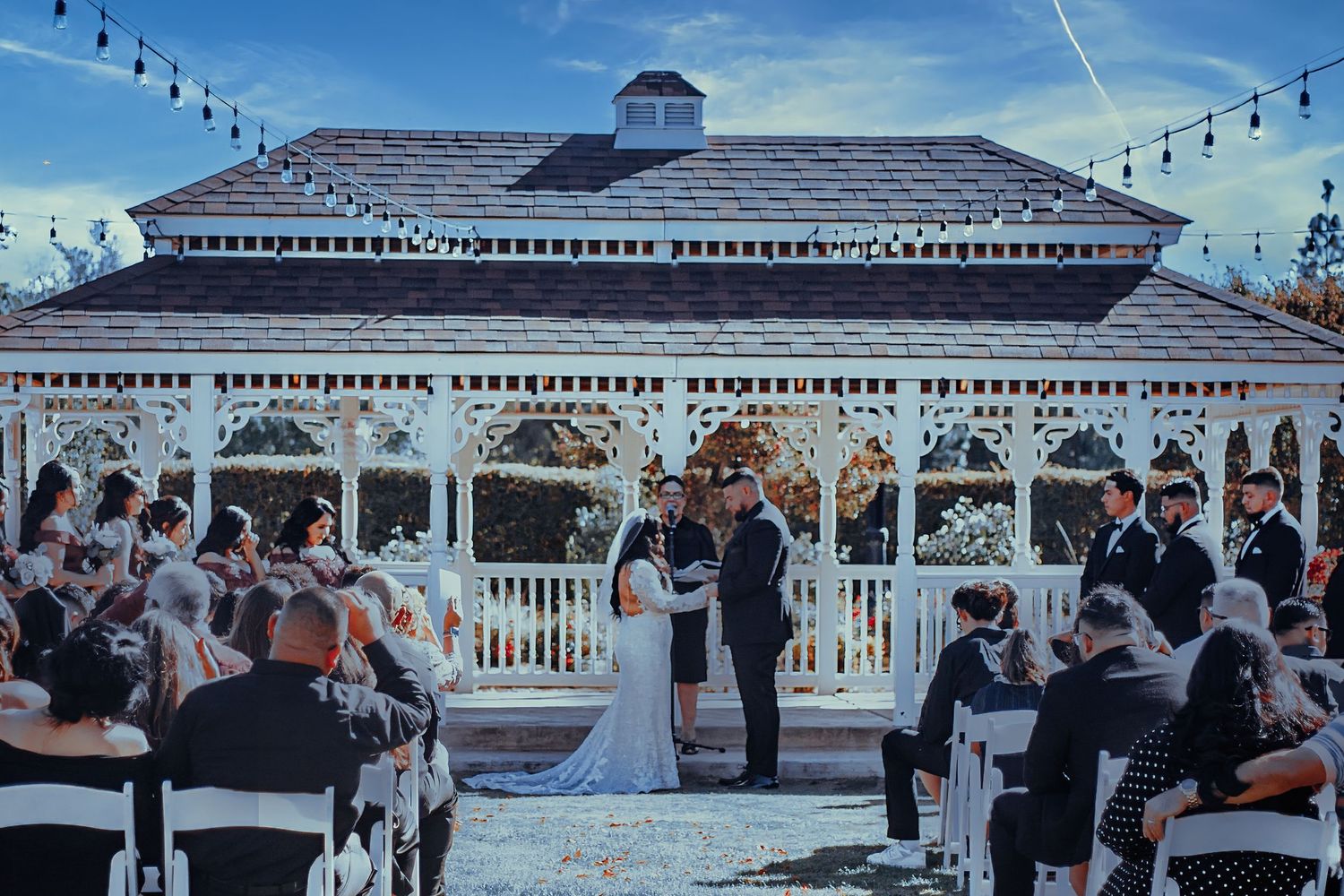 An outdoor wedding ceremony takes place under a white gazebo with string lights at dusk as guests watch from white chairs.